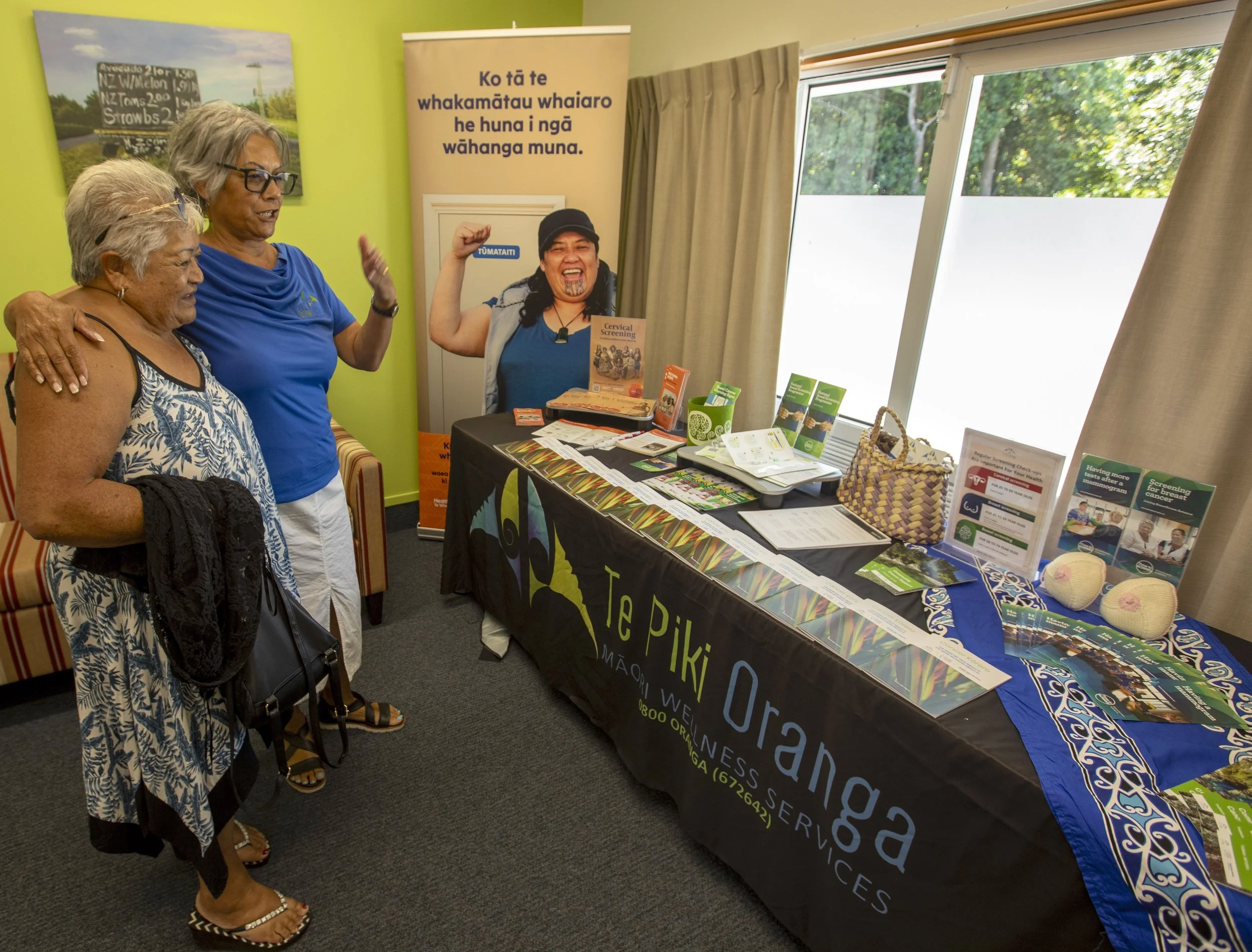 Two women and a baby sitting on a beach, with one woman holding the baby's foot and the other holding the baby. There are pieces of driftwood and logs around them.