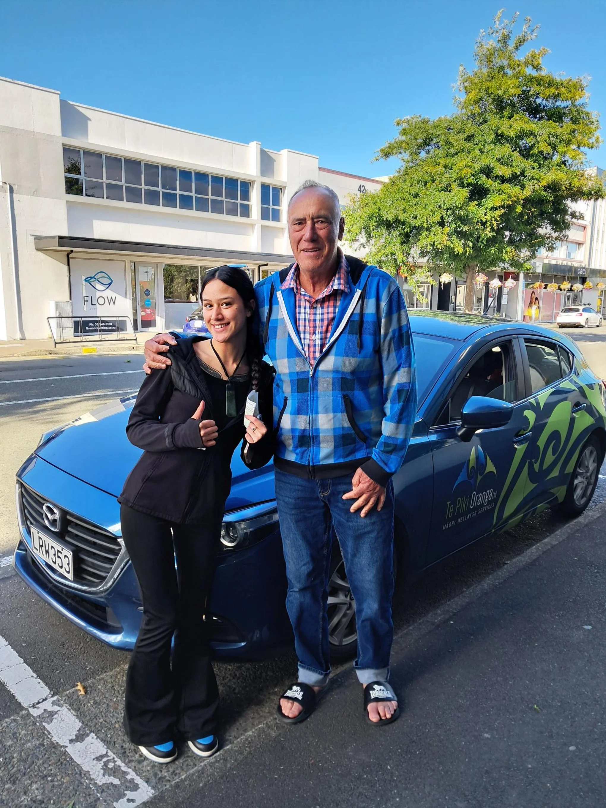 A young woman and an older man standing on a city street in front of a blue car. The young woman is giving a thumbs-up and smiling, while the older man has his arm around her. The background shows commercial buildings, trees, and a clear blue sky.