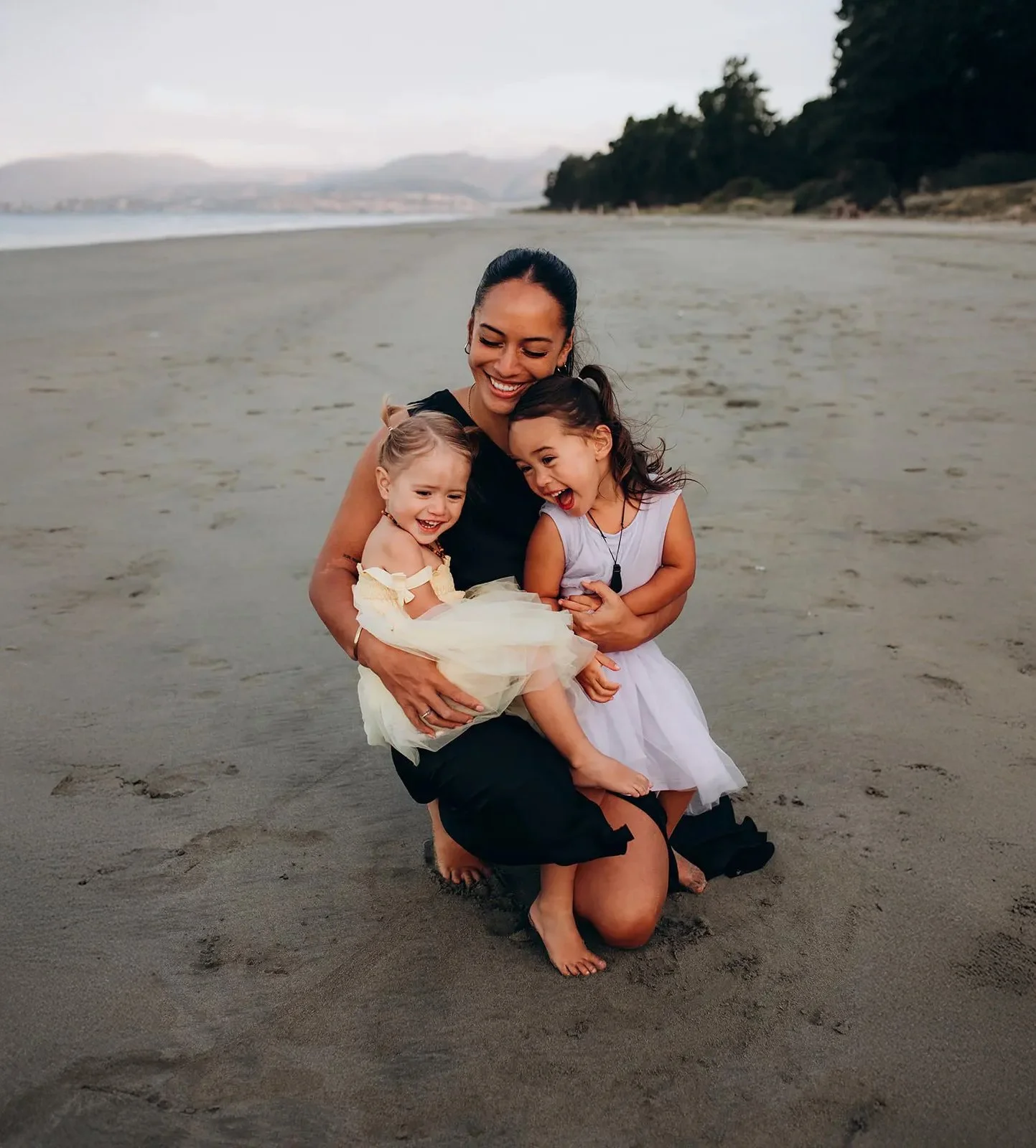 A woman and two young girls are happily playing and hugging each other on a sandy beach.
