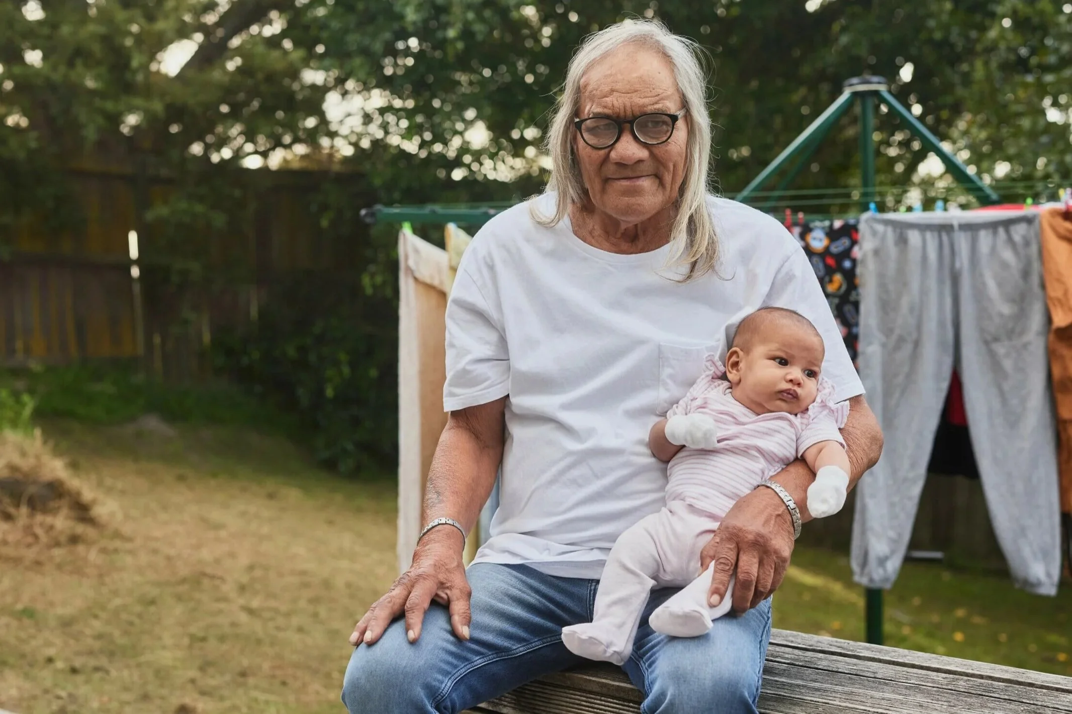 An elderly woman with glasses holding a baby on her lap in backyard with laundry hanging on drying rack.