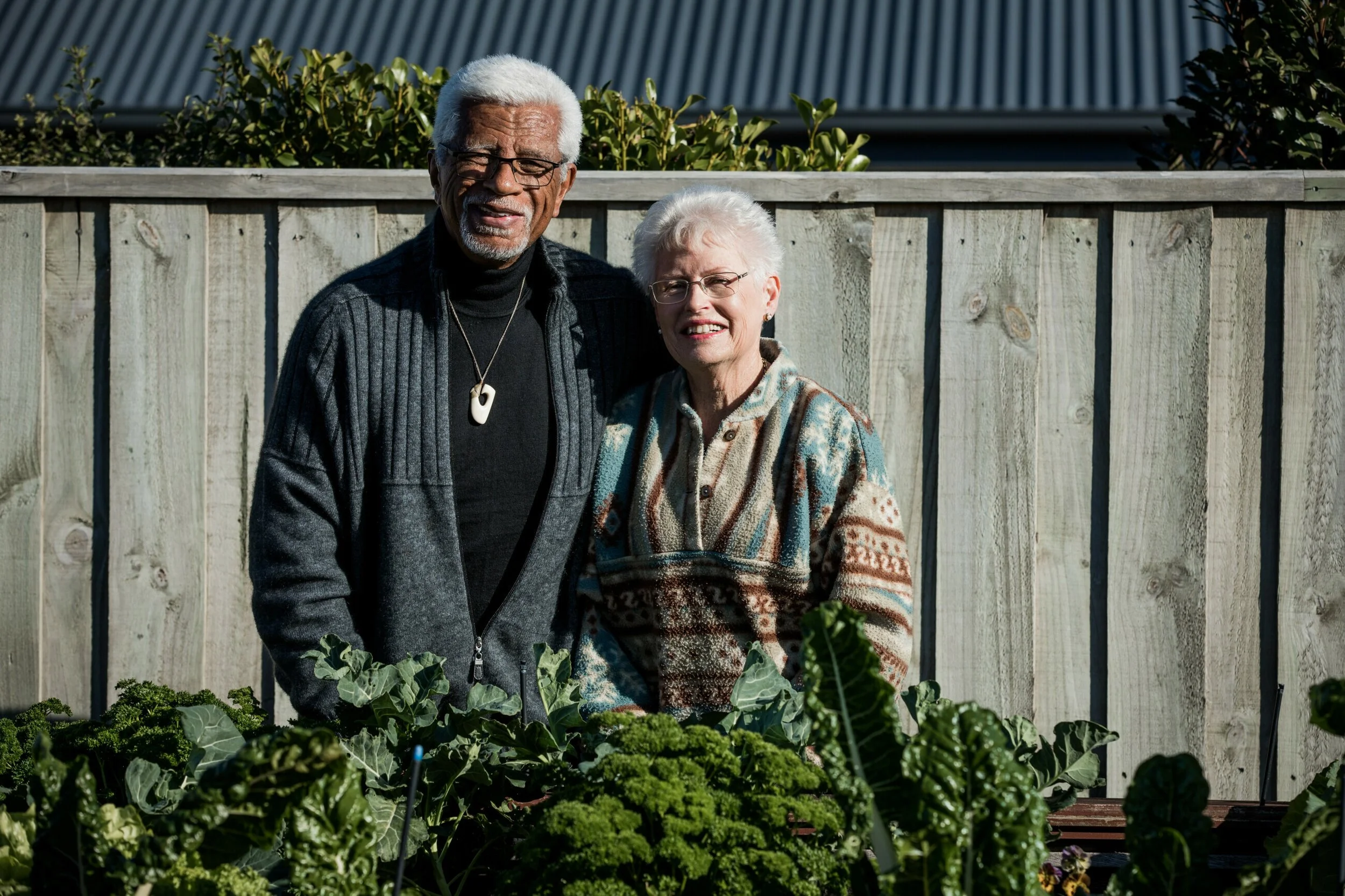An elderly couple standing outdoors in a garden with green plants, smiling at the camera, with a wooden fence in the background.