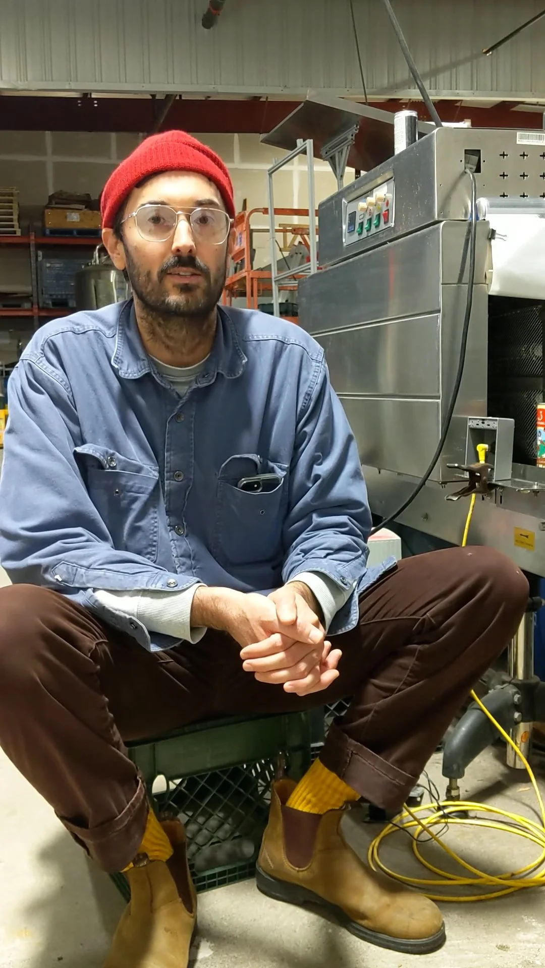 A man sitting cross-legged in a workshop next to a large industrial machine, wearing glasses, a red beanie, a denim shirt, brown pants, yellow socks, and tan work boots.