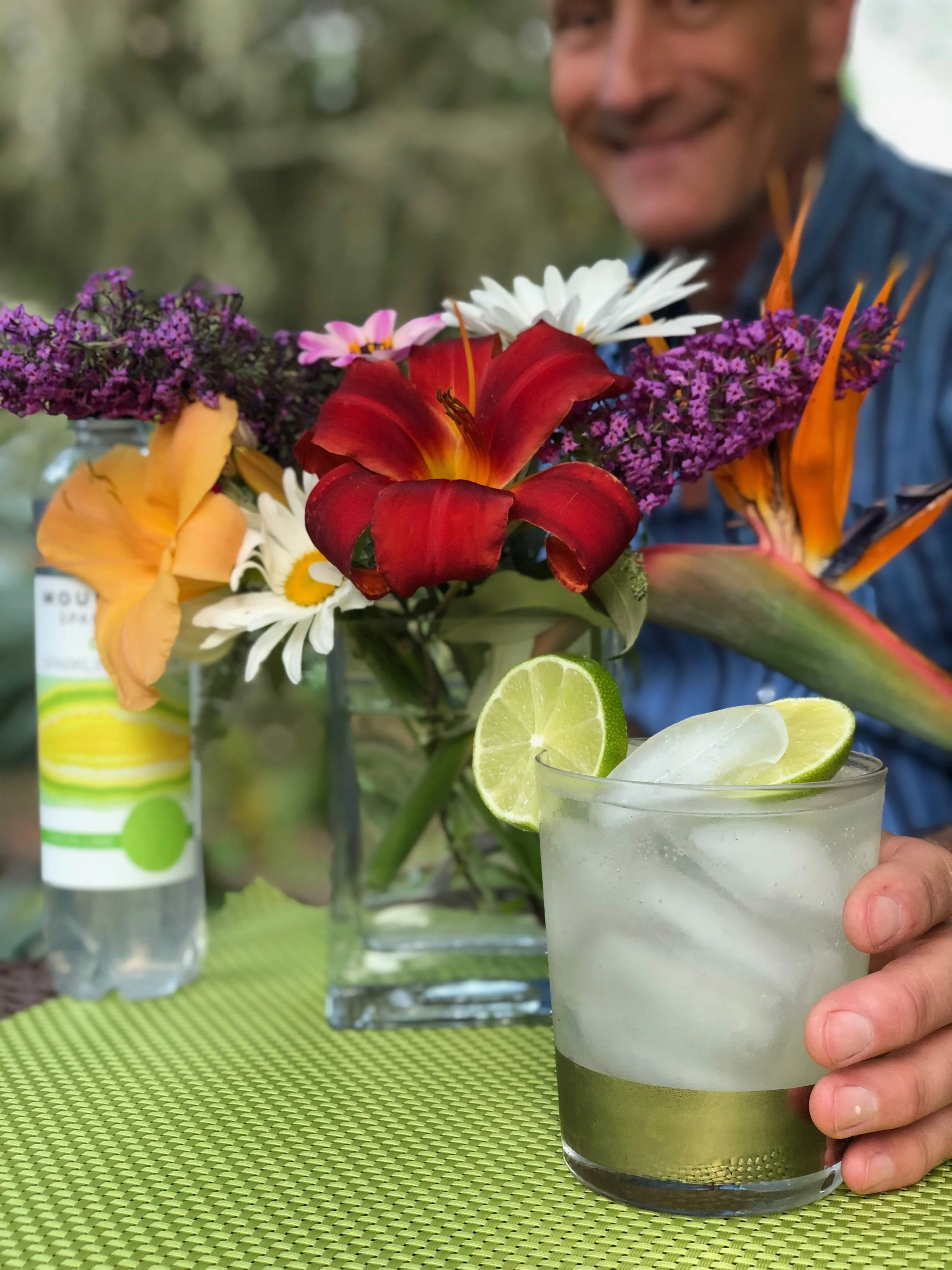 A person with a mustache smiling while holding a glass of limeade with ice and lime slices, with a bouquet of colorful flowers in the background on a green table mat.