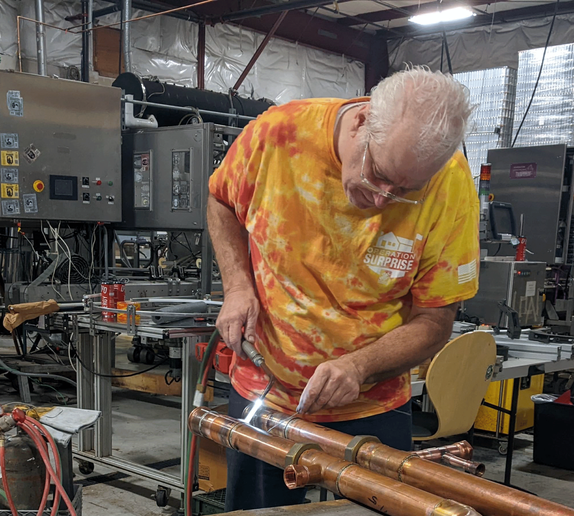 An elderly man with white hair and glasses working on copper pipes in a workshop.
