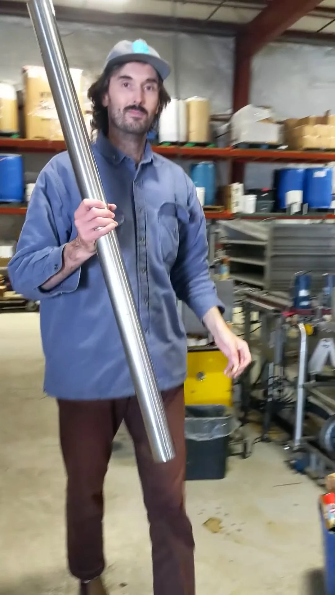 Man standing in a workshop holding a large metal pipe, surrounded by industrial supplies and equipment.
