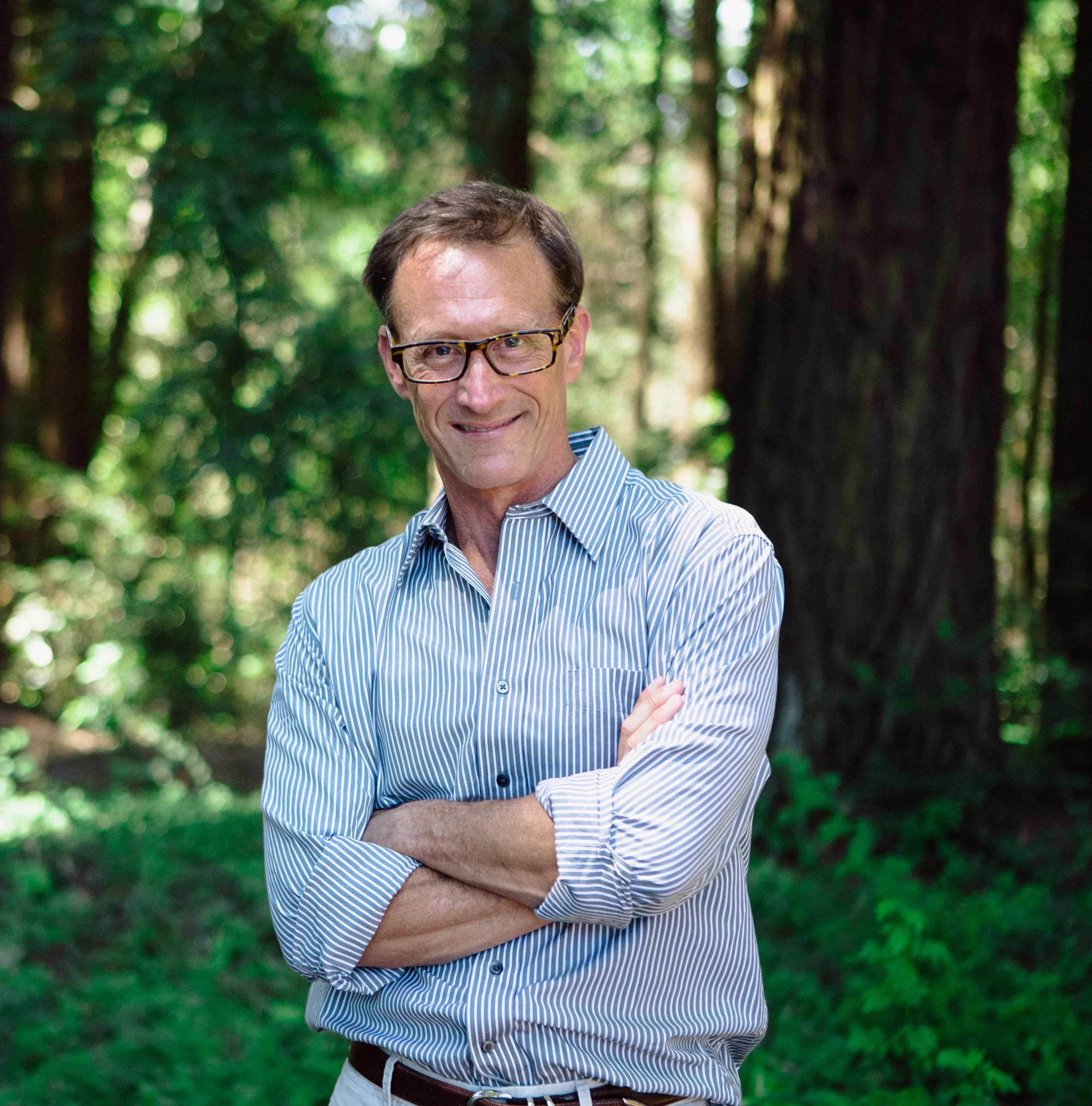 A man with glasses and a striped shirt standing with arms crossed in a forest with tall trees and green foliage.