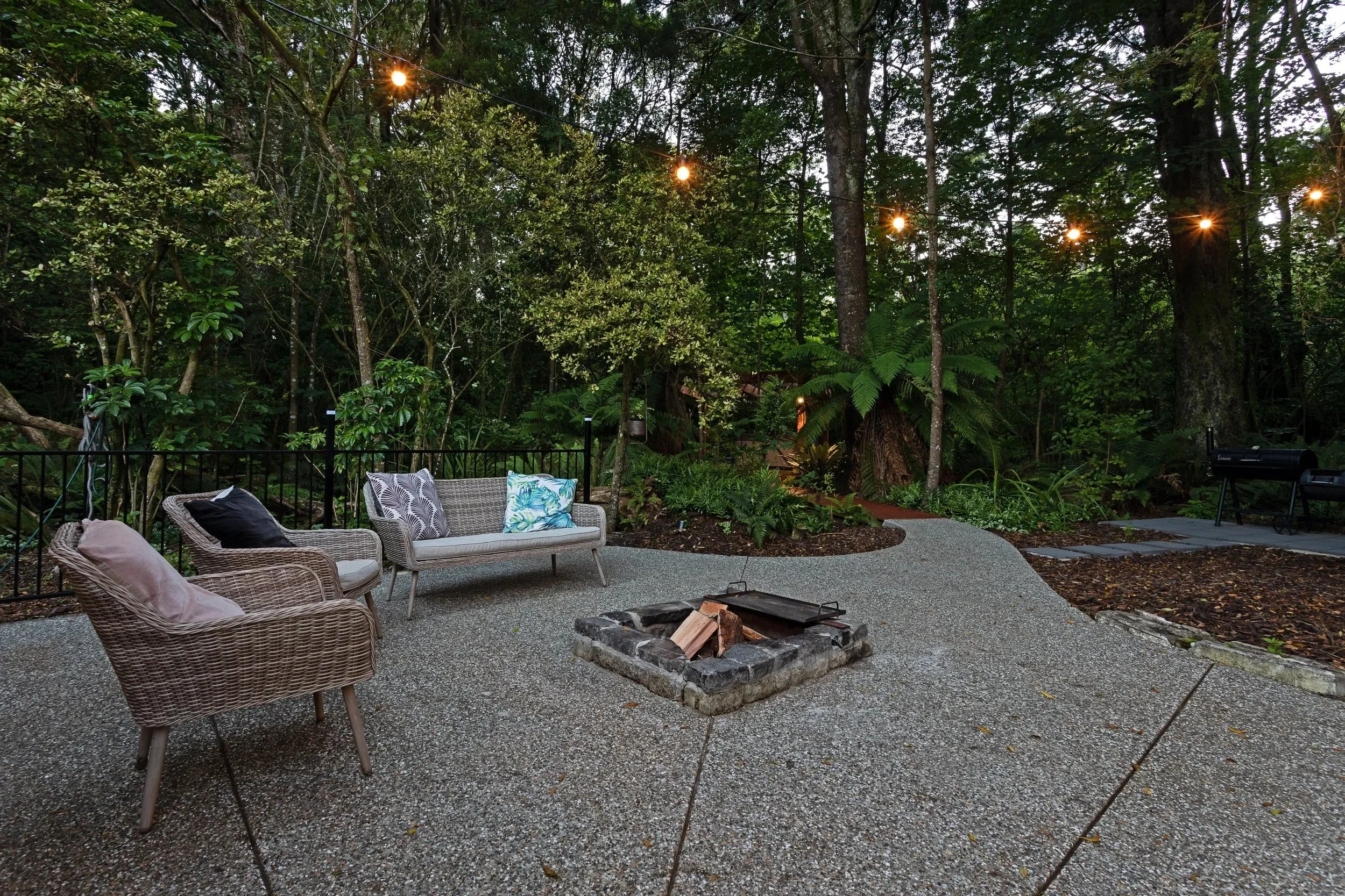 Outdoor patio at dusk with wicker and fabric chairs with pillows, a stone firepit with wood, surrounded by nature.
