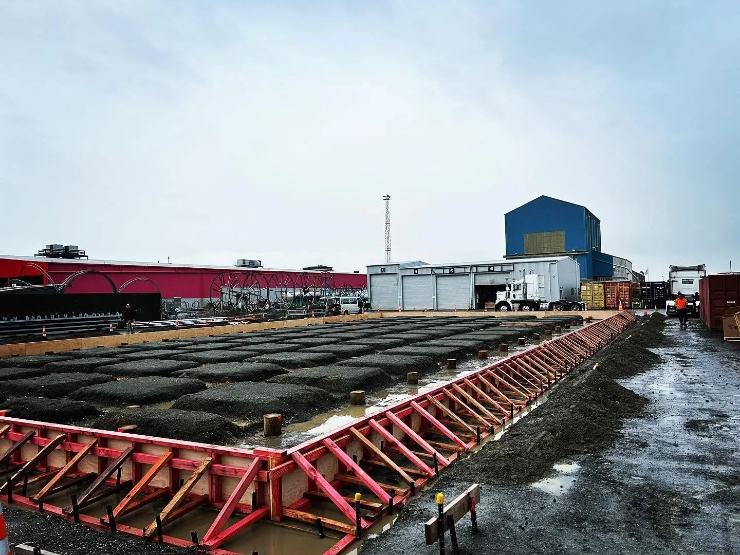Construction site with a large area of poured concrete, red formwork, and industrial buildings in the background on a cloudy day.