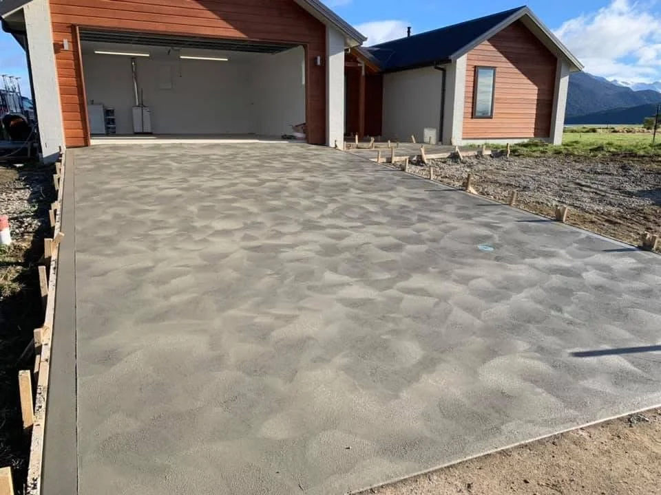 Newly poured concrete driveway leading to a house with an open garage and a wooden exterior, set against a backdrop of mountains and blue sky.