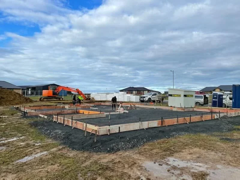 Construction site with foundation framing, construction workers, construction equipment, and portable toilets, in a residential area under cloudy sky.