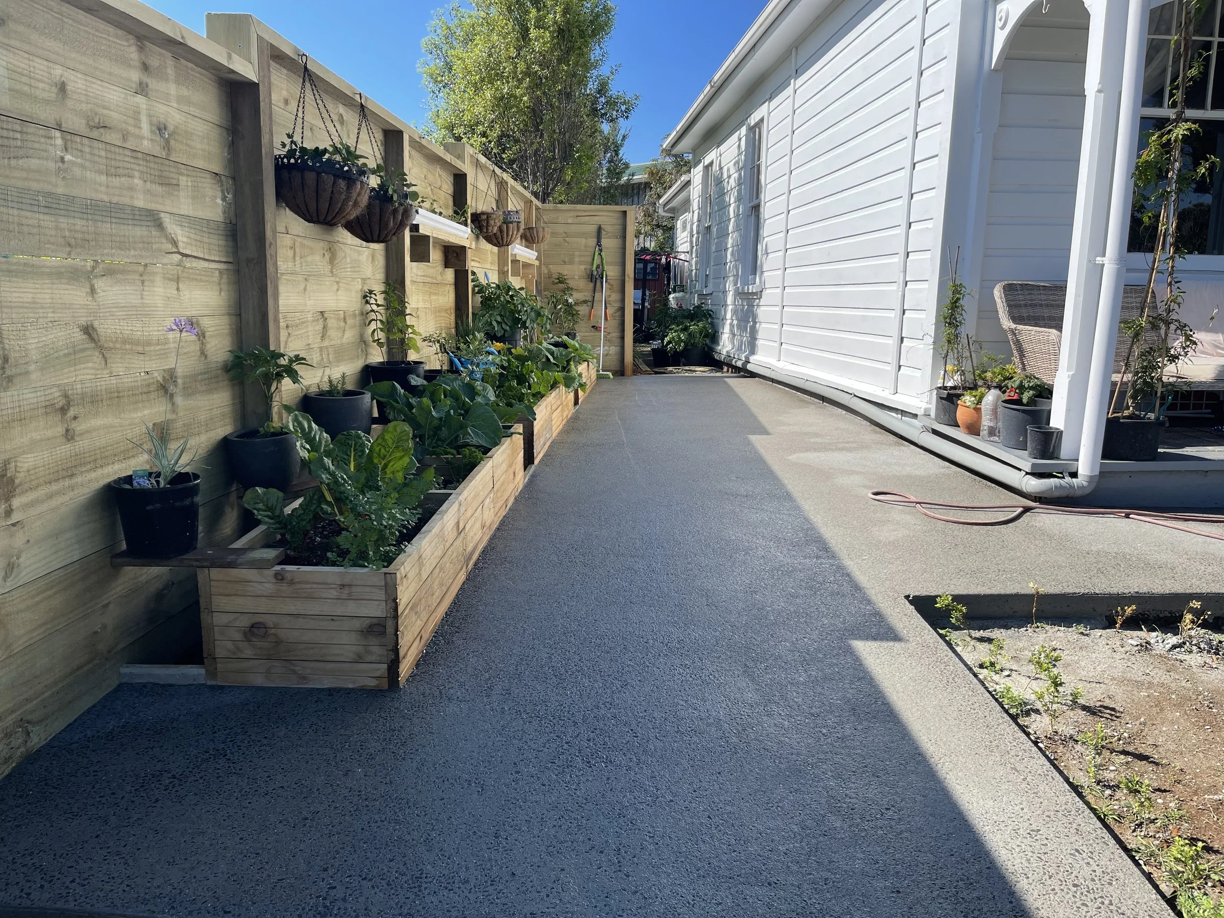 A backyard patio with a wooden fence, potted plants, and a white house with a patio area, on a sunny day.