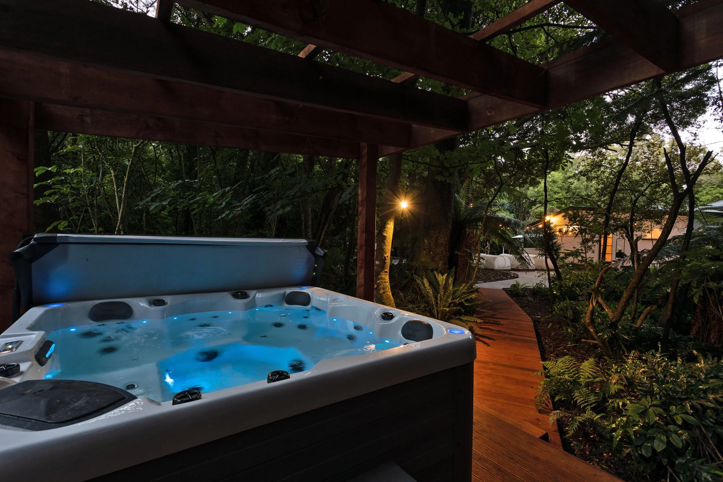 An outdoor hot tub with blue LED lights in a wooden deck covered patio, surrounded by lush green trees and plants, with warm outdoor lighting in the evening.