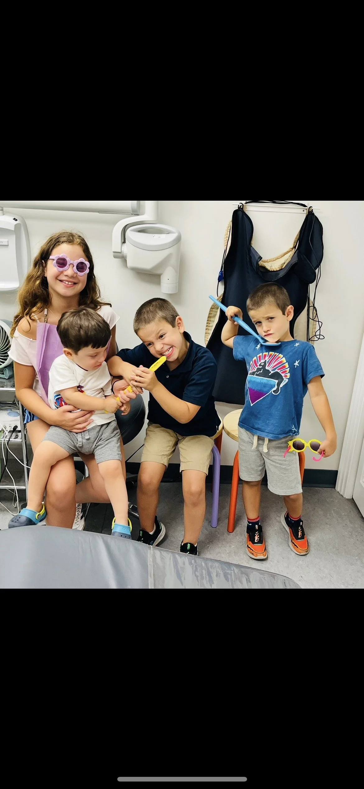 Four children having fun at a doctor’s office, with three sitting and one being held, wearing colorful sunglasses and holding toy accessories.