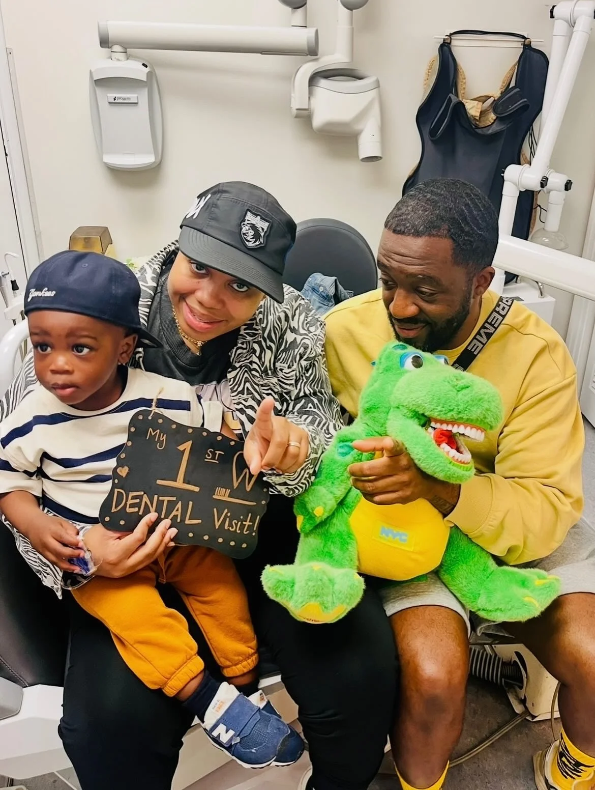 A woman, a young boy, and a man sitting in a dental office, celebrating the child's first dental visit. The woman holds a sign that says 'My 1st Dental Visit!' The man is holding a green stuffed dinosaur toy, and they are all smiling.