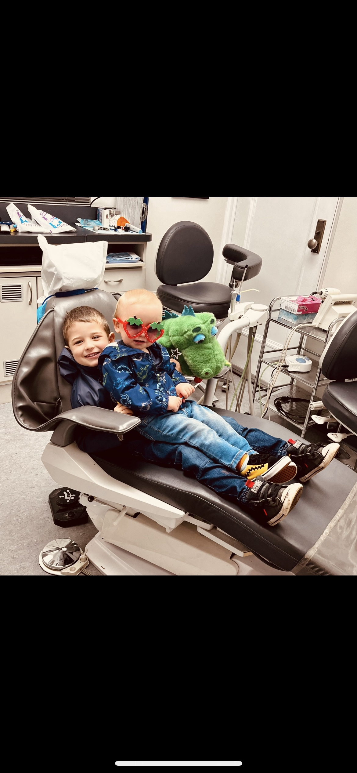 Two children, one with glasses and the other smiling, sitting on a medical examination table in a doctor's office, holding plush dragon toys, with medical equipment in the background.