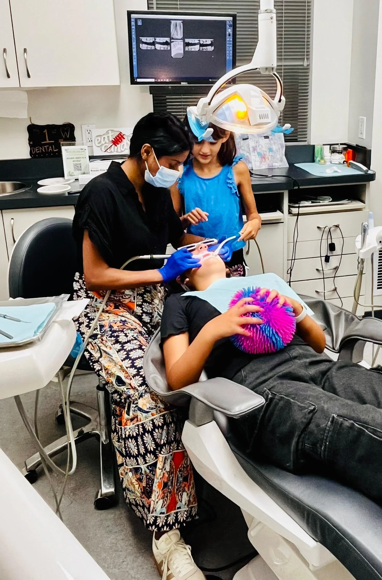 A child receiving dental treatment from a dentist at a dental clinic, with a dental assistant nearby and a monitor displaying dental X-rays in the background.
