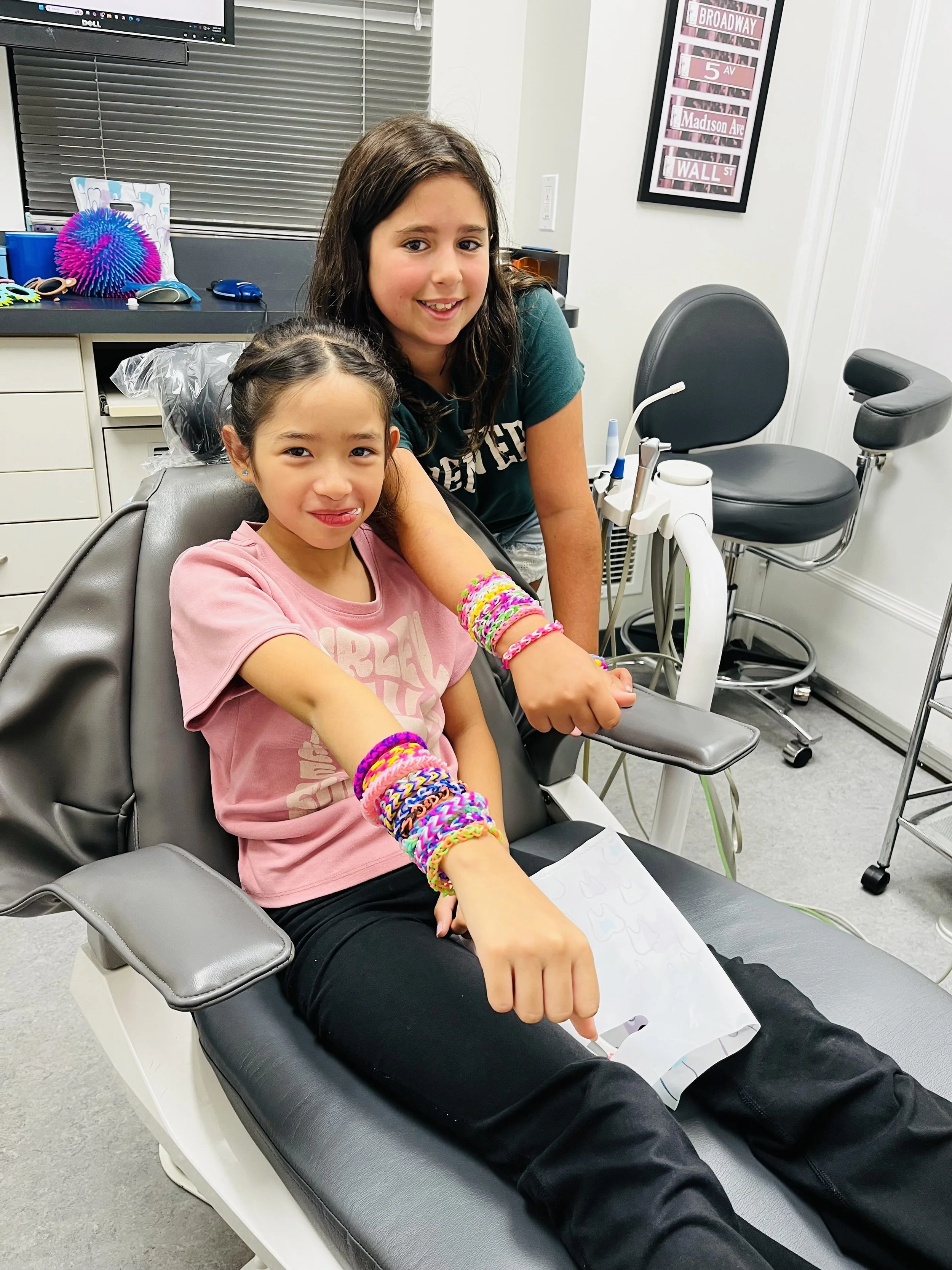 A young girl is seated in a dentist's chair with her arm extended, showing colorful rubber band bracelets. An older girl stands beside her, both smiling. The setting is a dental clinic with dental equipment and colorful decor.