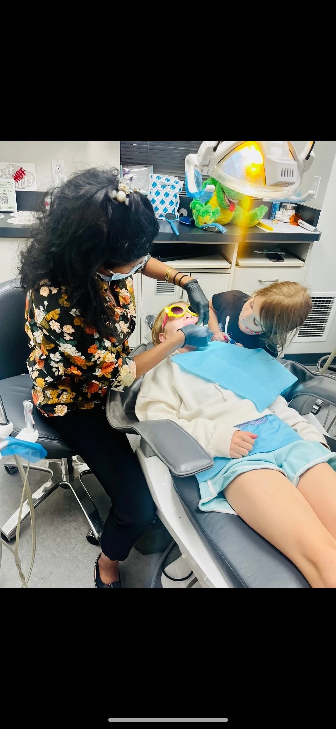 A young girl wearing sunglasses sitting in a dental chair as a dentist examines her teeth, with another girl leaning over her smiling, in a brightly lit dental office.