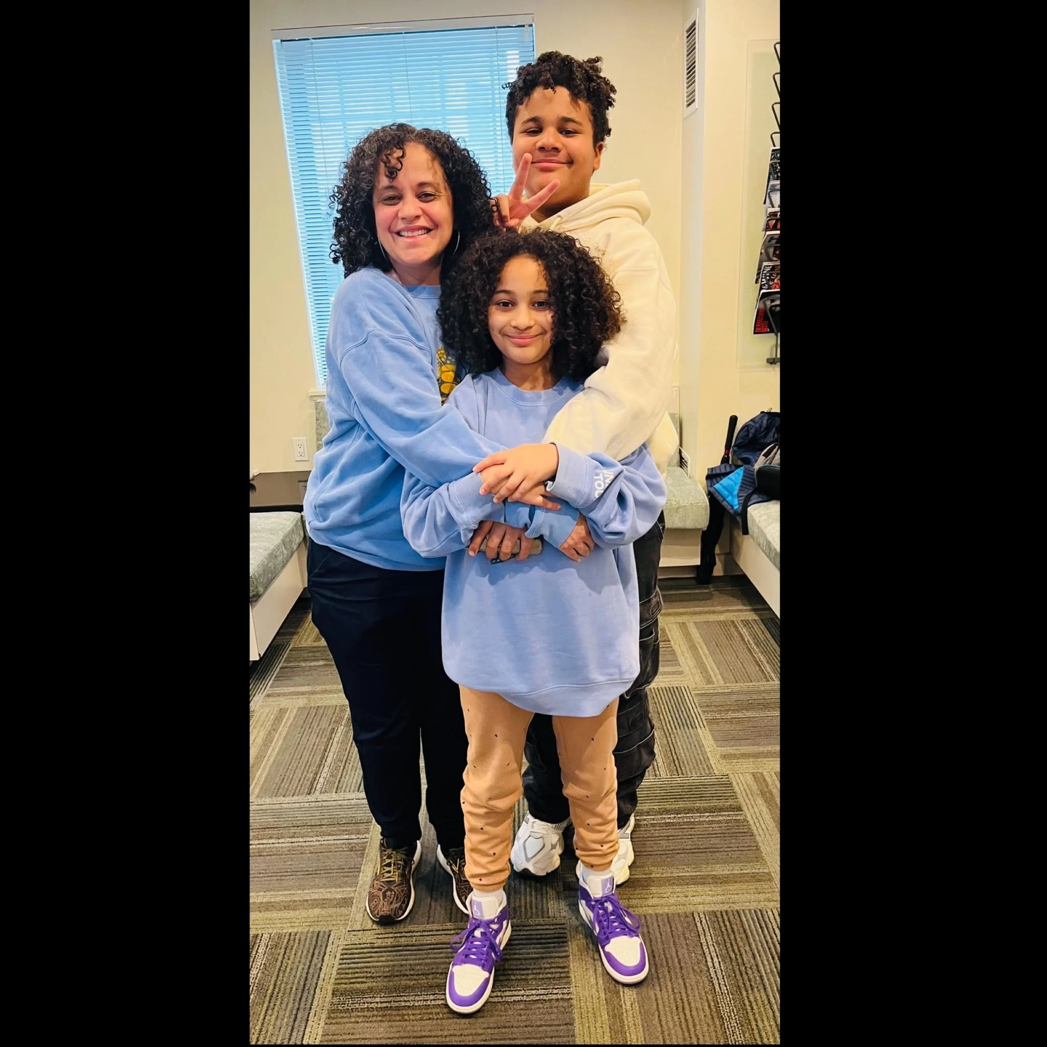 A woman and two children smiling and hugging each other in a room with a window, benches, and backpacks in the background.