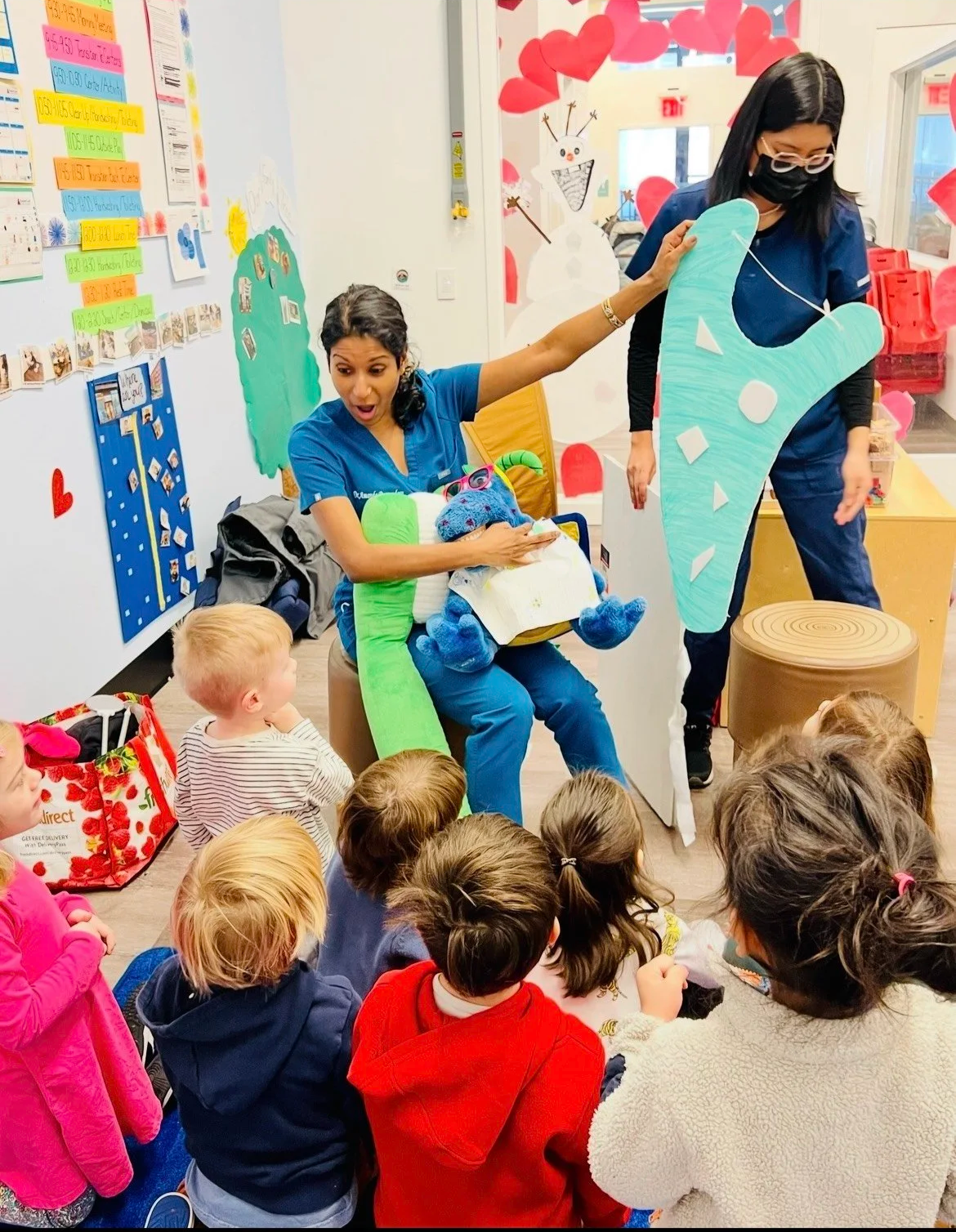 Two women dressed as a green sock and a slice of cake are performing puppet show for children on the floor in a colorful classroom decorated with hearts and snowman artwork.