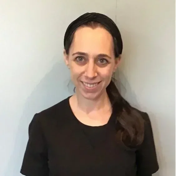 A woman with long dark hair in a ponytail, wearing a black top, smiling and standing against a plain light-colored wall.