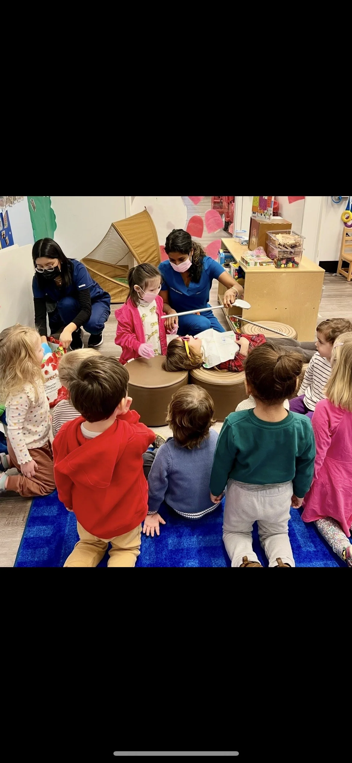 Children and adults gathered around as a person performs a magic trick with a prop in a classroom or play area, with shelves of toys and educational materials in the background.