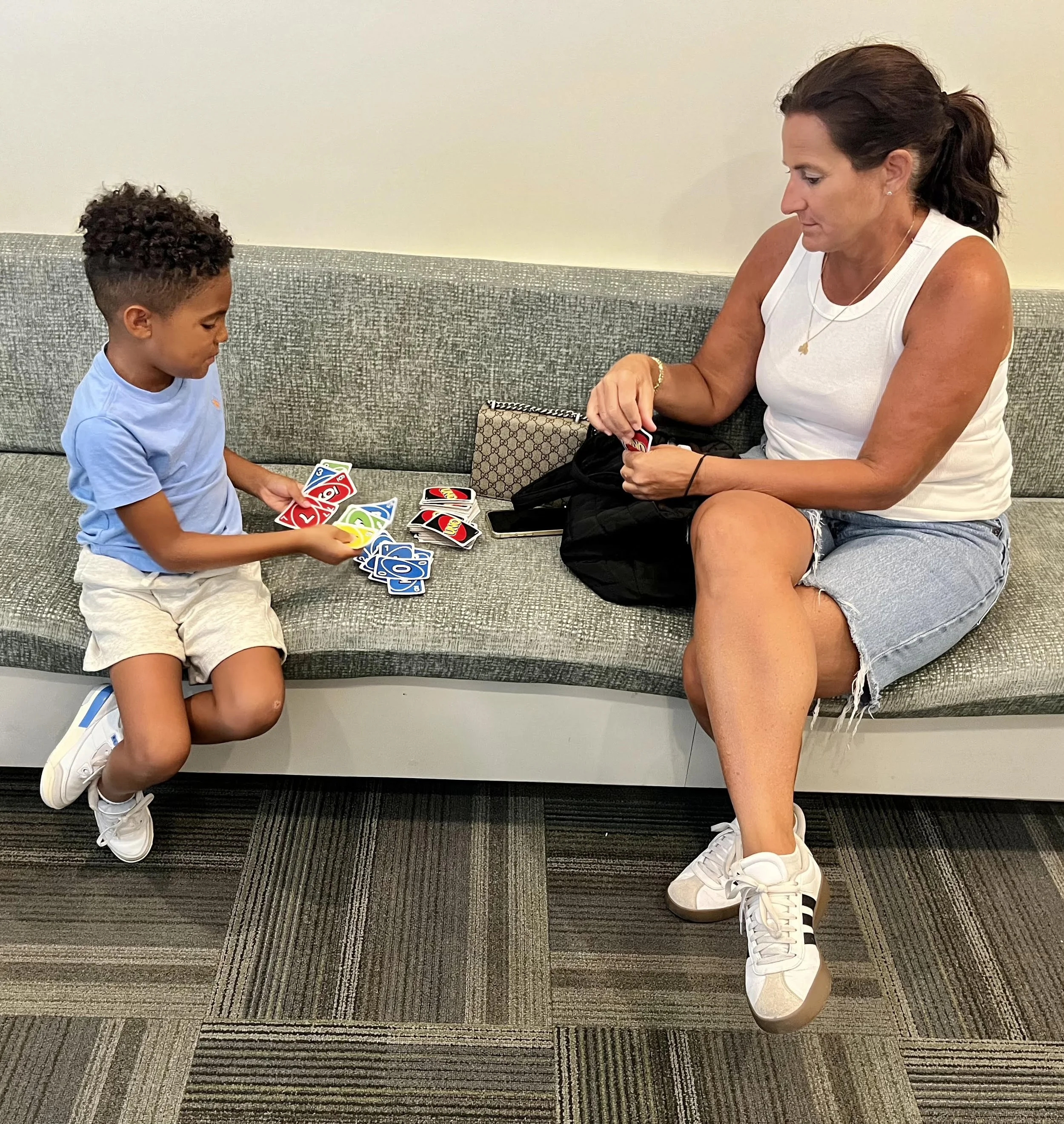 A woman and a young boy are sitting on a gray bench, playing UNO with the cards spread out in front of them. The woman is sorting her cards while the boy is holding multiple cards and looking at them. The woman has long dark hair tied back, and is wearing a white sleeveless top and ripped denim shorts. The boy has short curly hair and is wearing a light blue T-shirt and beige shorts. There is a designer purse and a cellphone on the bench next to them.