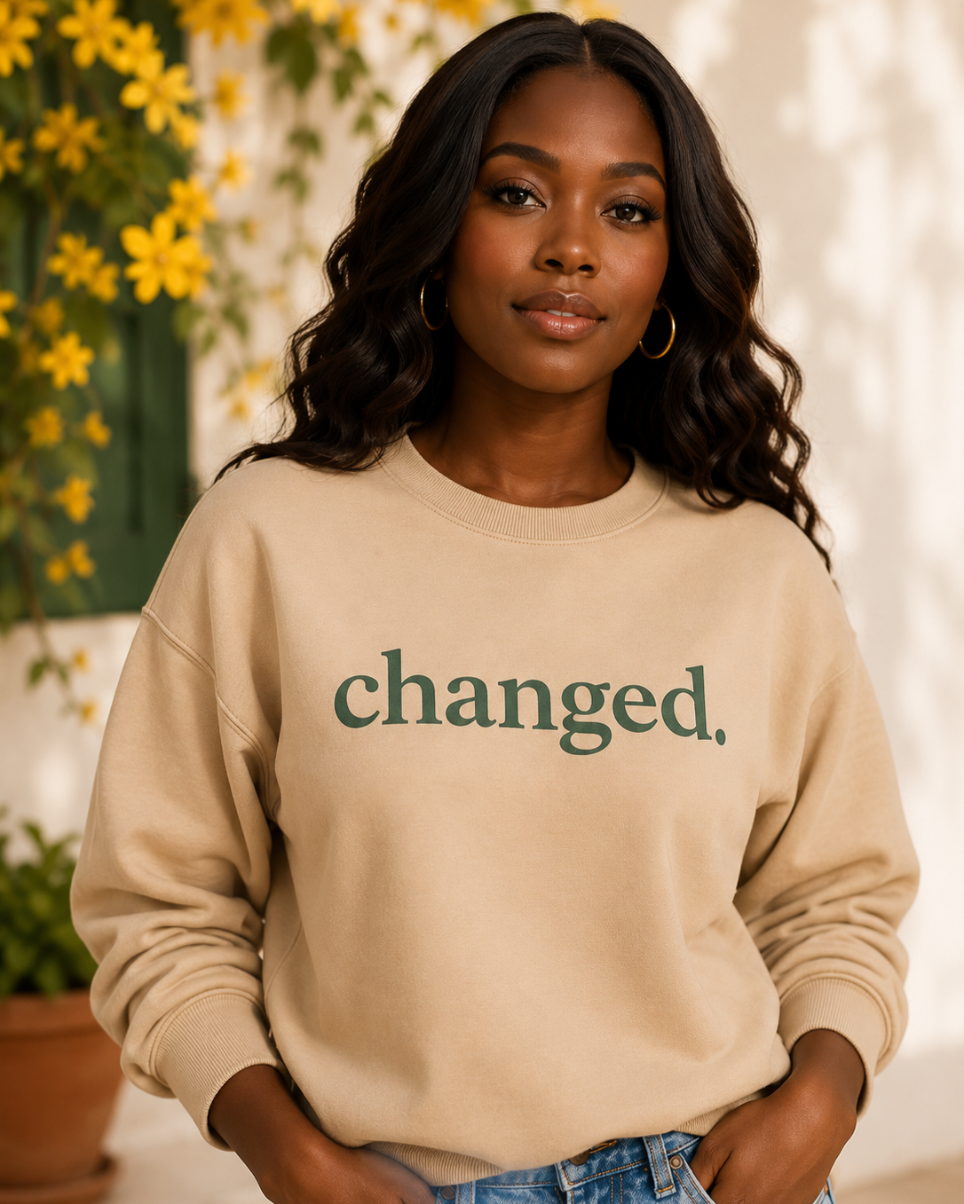 Woman with dark wavy hair wearing a beige sweatshirt that says 'changed.' in green, standing outdoors with yellow flowers in the background.