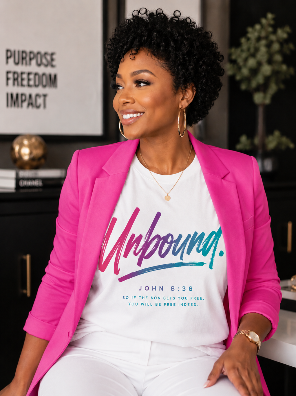 Smiling woman with curly black hair wearing a pink blazer and graphic t-shirt with a Bible verse, sitting indoors with a black shelf, a plant, and a framed text in the background.
