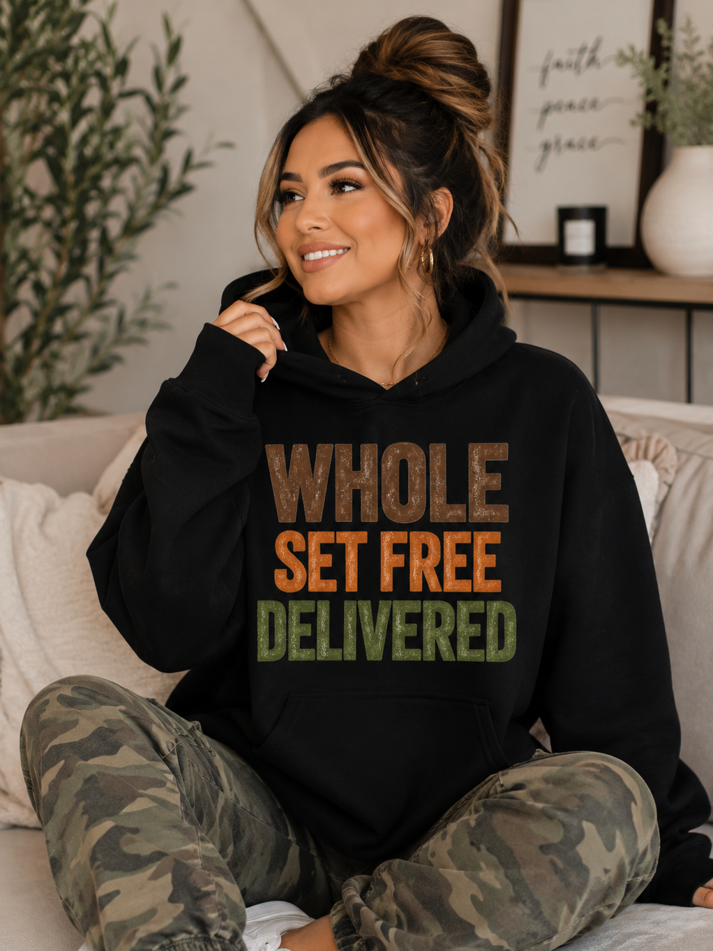 A smiling woman sitting cross-legged on a sofa, wearing a black hoodie that says 'WHOLE SET FREE DELIVERED' in brown, orange, and green text, with camouflage pants and hoop earrings, in a cozy, decorated living room.