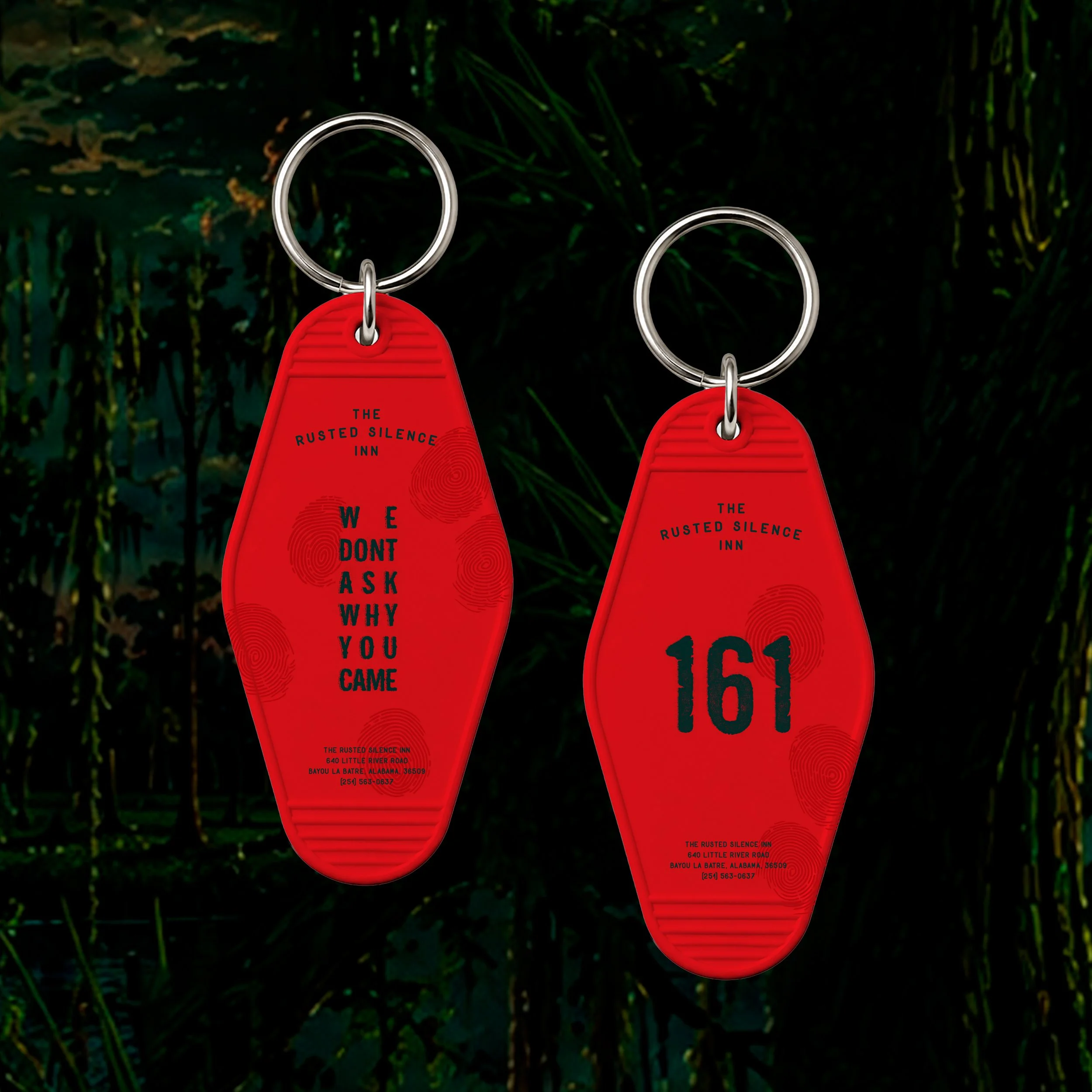 Close-up of two red keychains with black text hanging from silver key rings against a dark, green, leafy background. The keychains include a message stating, "WE DONT ASK WHY YOU CAME" and an address for The Rusted Silence Inn, located at 640 Little River Road, Bayou La Batre, Alabama, with a phone number.