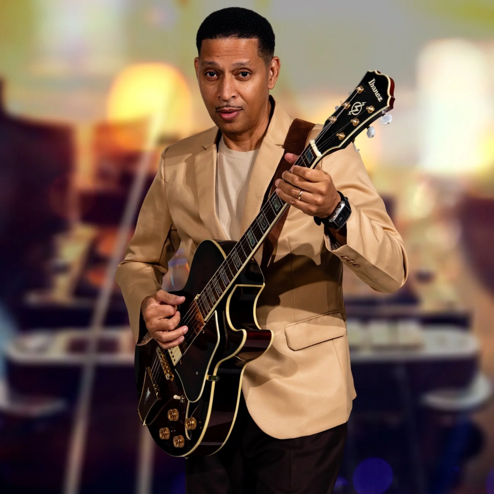 Man in tan suit plays brown guitar in front of blurred background.