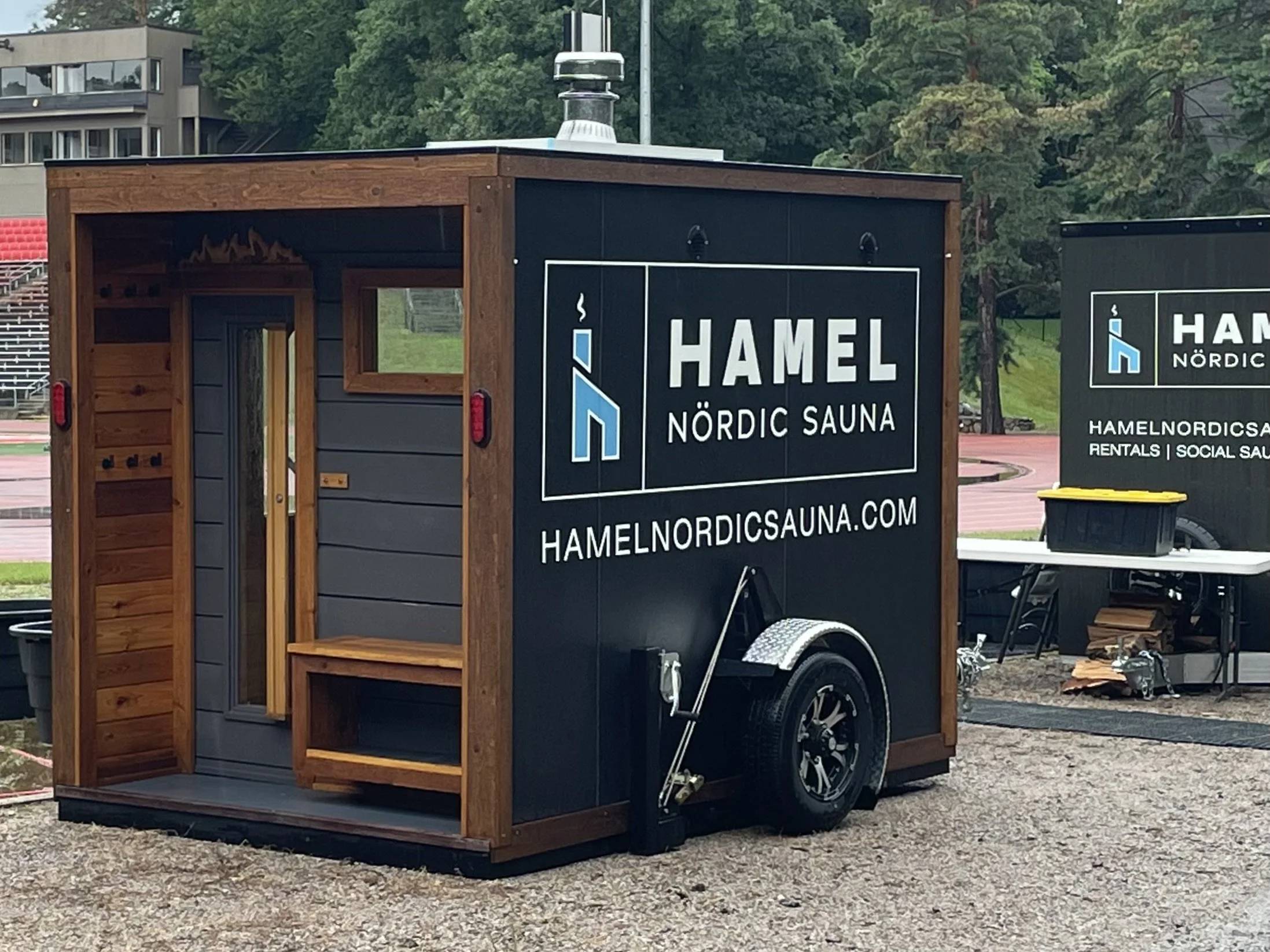 A small mobile sauna trailer with a black exterior and wooden accents, displaying the words 'Hamel Nordic Sauna' and a website URL, parked outdoors on a gravel surface. In the background sits another Hamel Nordic Sauna mobile unit.