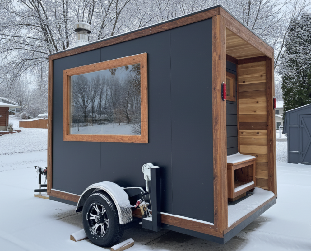 Custom-built mobile sauna with dark exterior, wooden trim, and a large window, set on a snowy landscape.