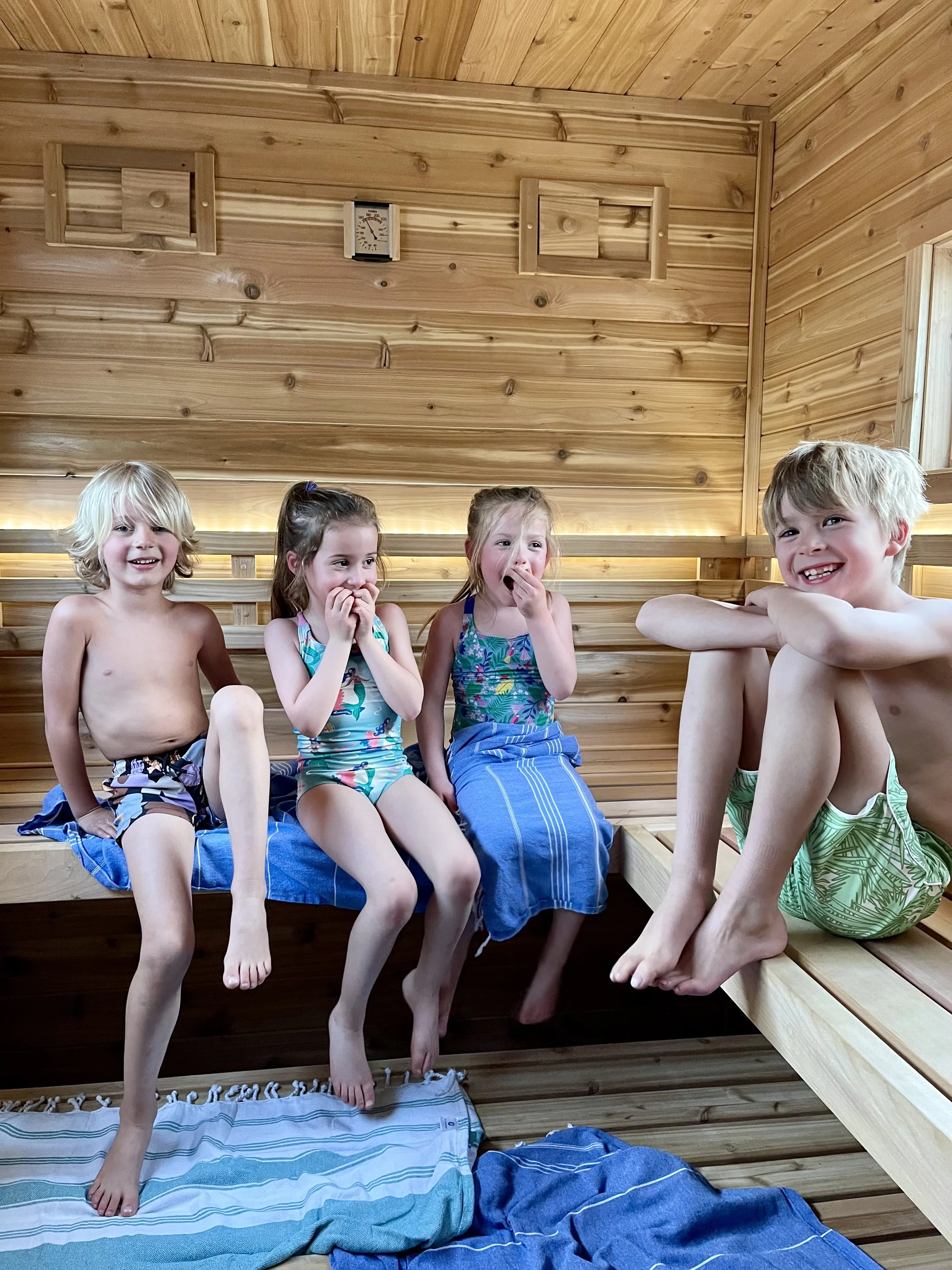 Four children sitting on the edge of a wooden sauna with their feet in the sauna and smiling or laughing.