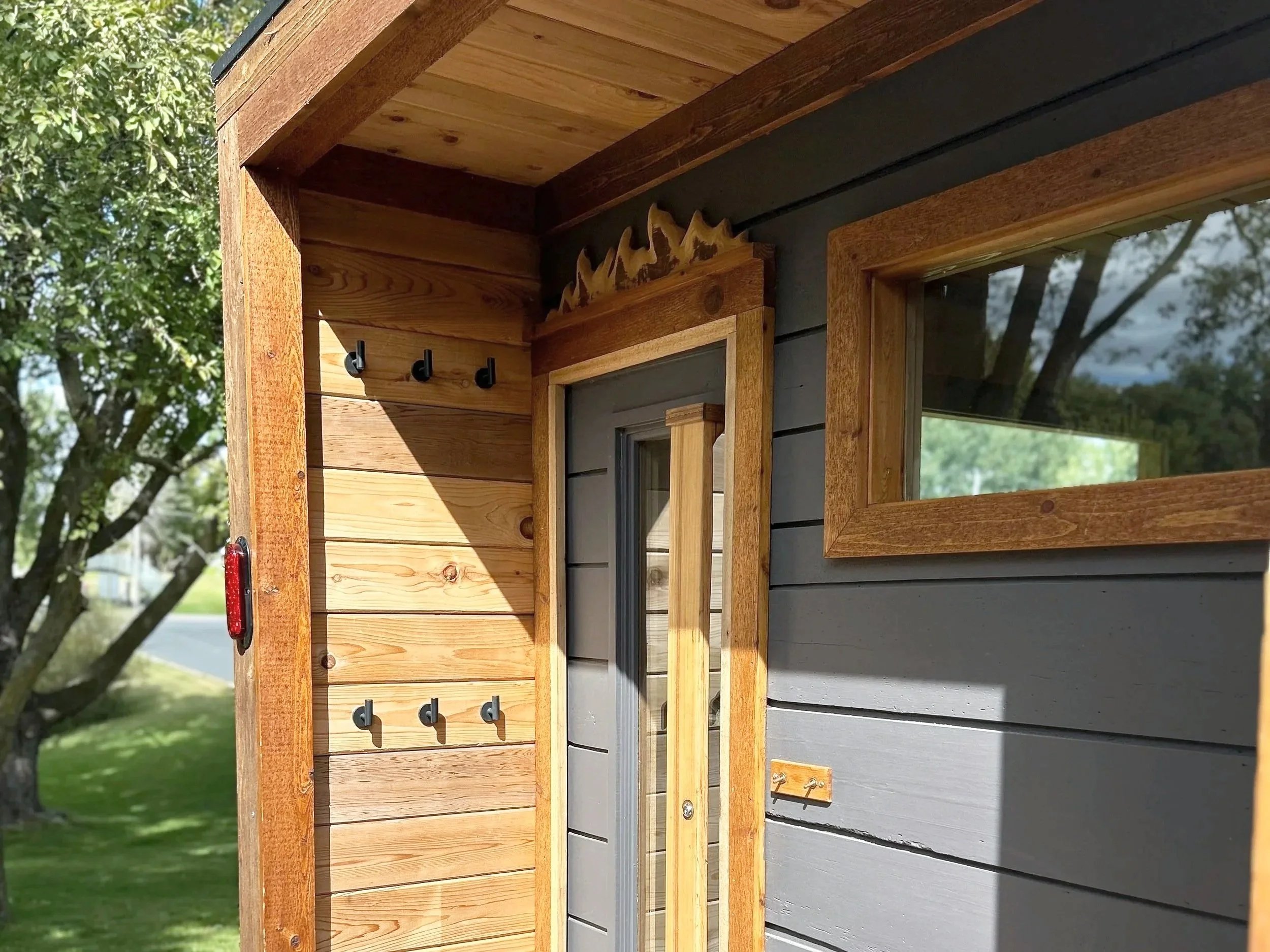 Close up of sauna exterior threshold, featuring six hooks, a door with a decorative wooden flame detail, and a transom window.