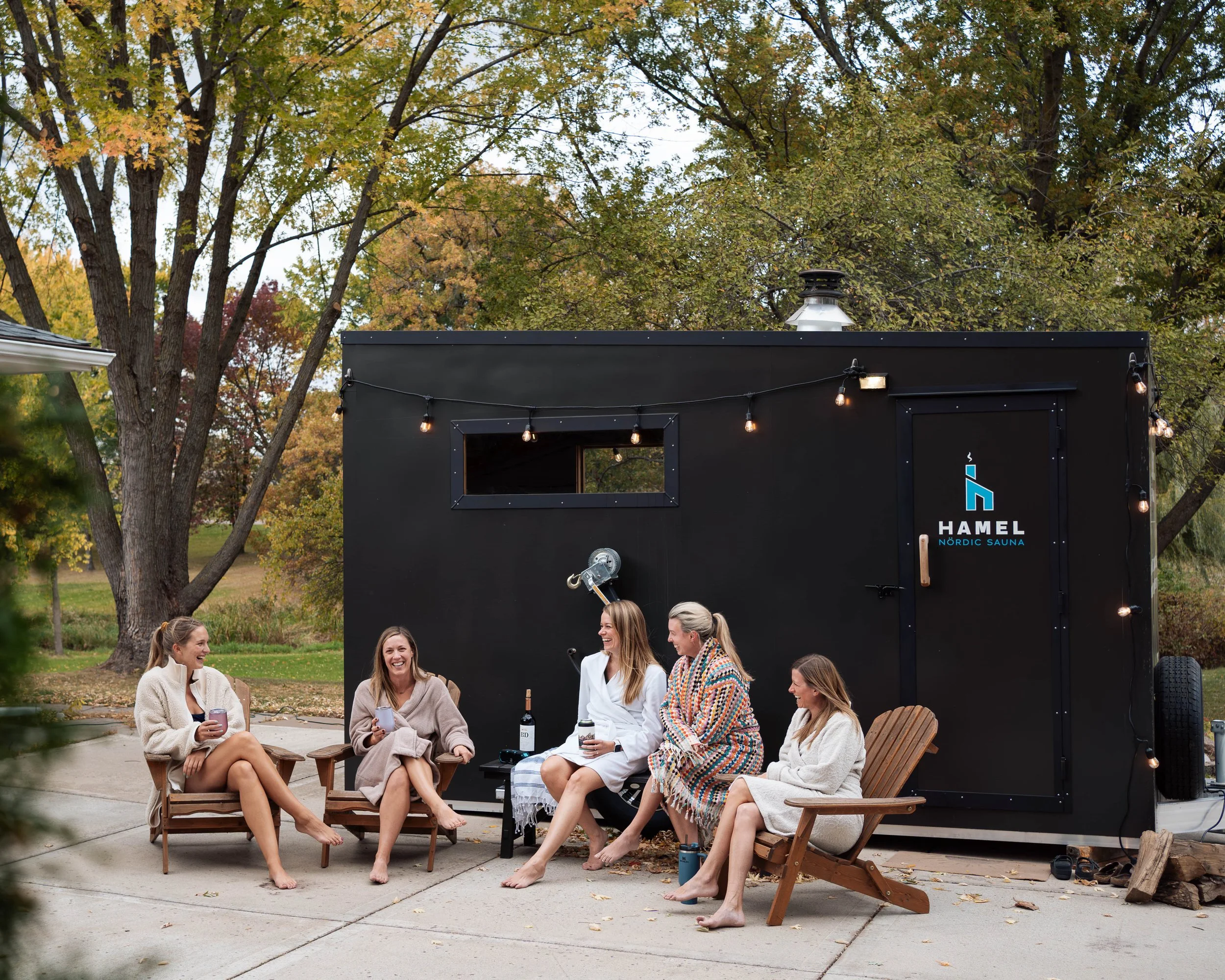 Five women sitting outside a black sauna trailer with the Hamel Nordic Sauna logo on the door, enjoying conversation, surrounded by trees with colorful fall leaves.