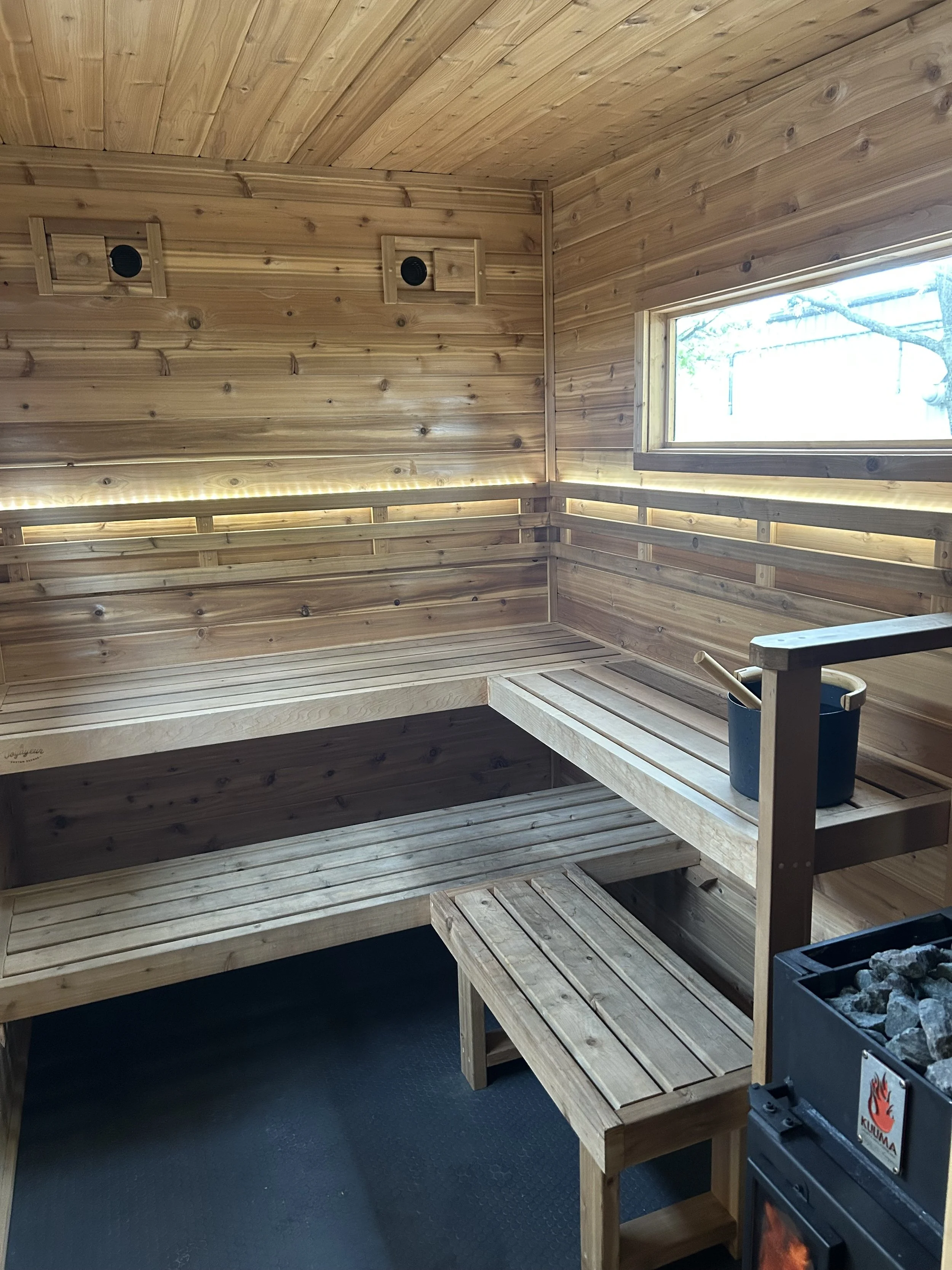 Interior of a wooden sauna room with benches, a window, and a Kuuma sauna stove with rocks.