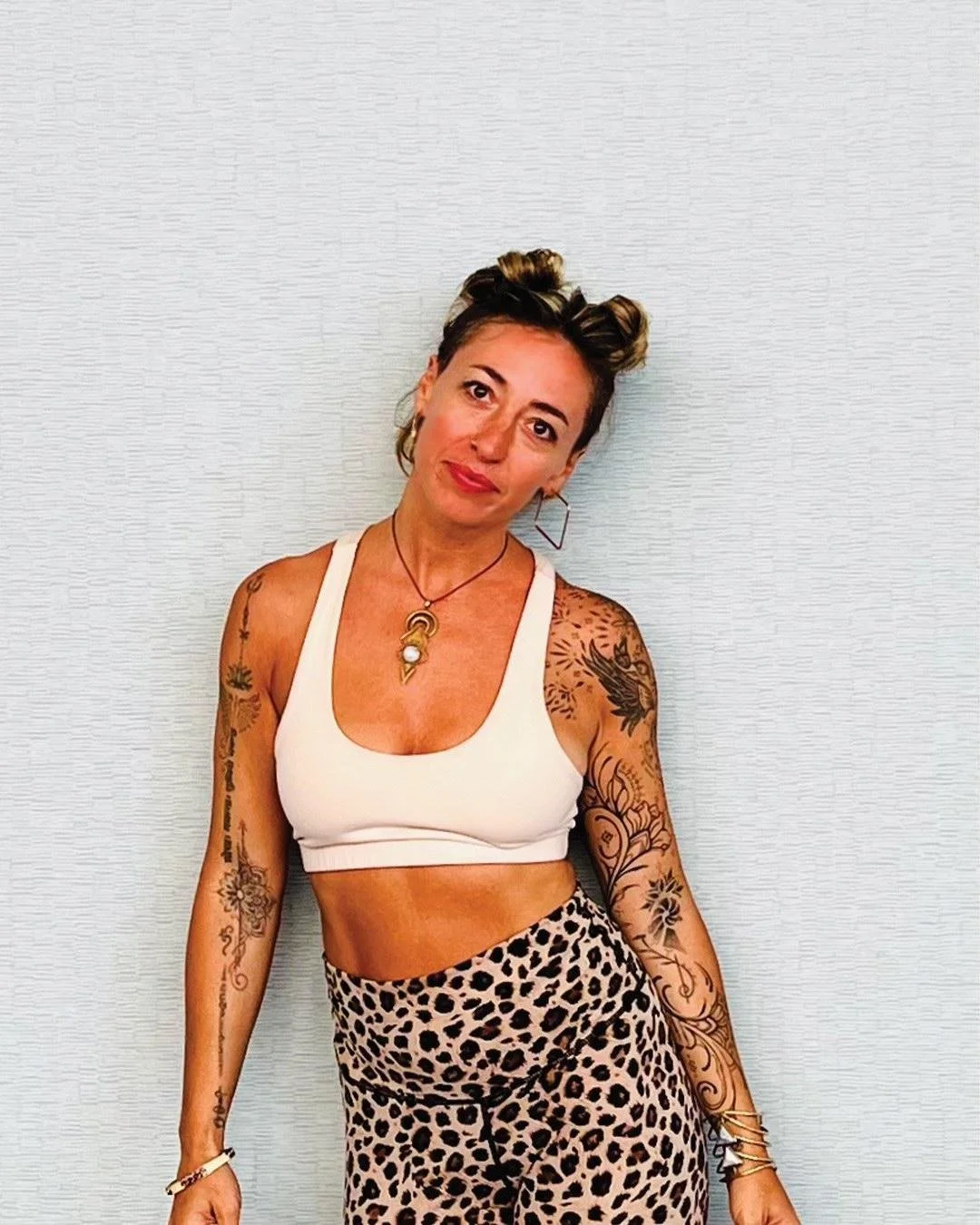 Dani Capra - A woman with tattoos on her arms and short, styled hair, wearing a white crop top and leopard print pants, standing against a plain wall.