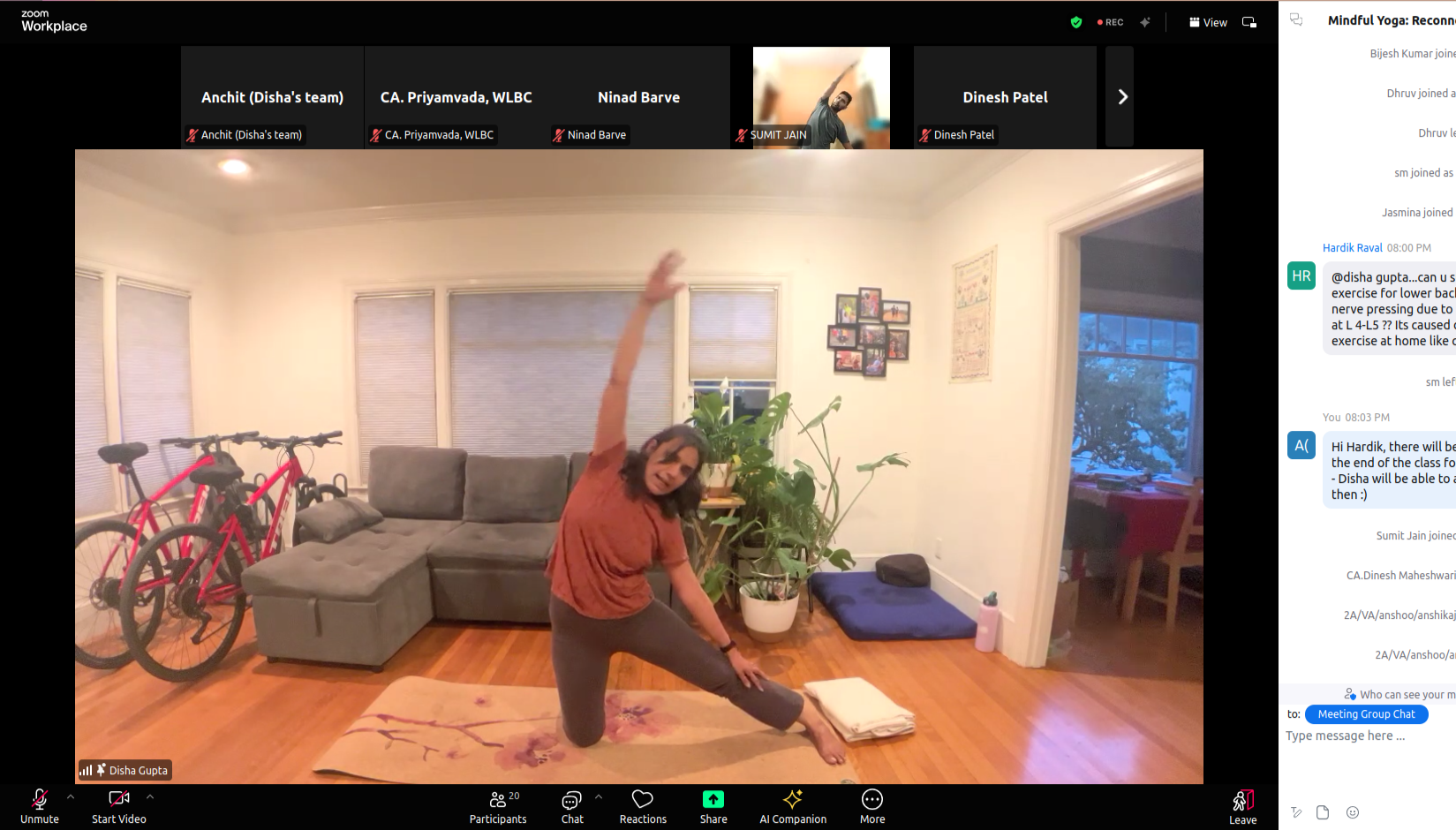 Woman practicing yoga in a living room, stretching with one arm raised above her head, on a pink and purple mat, surrounded by some houseplants, a gray sofa, and bicycles in the background.