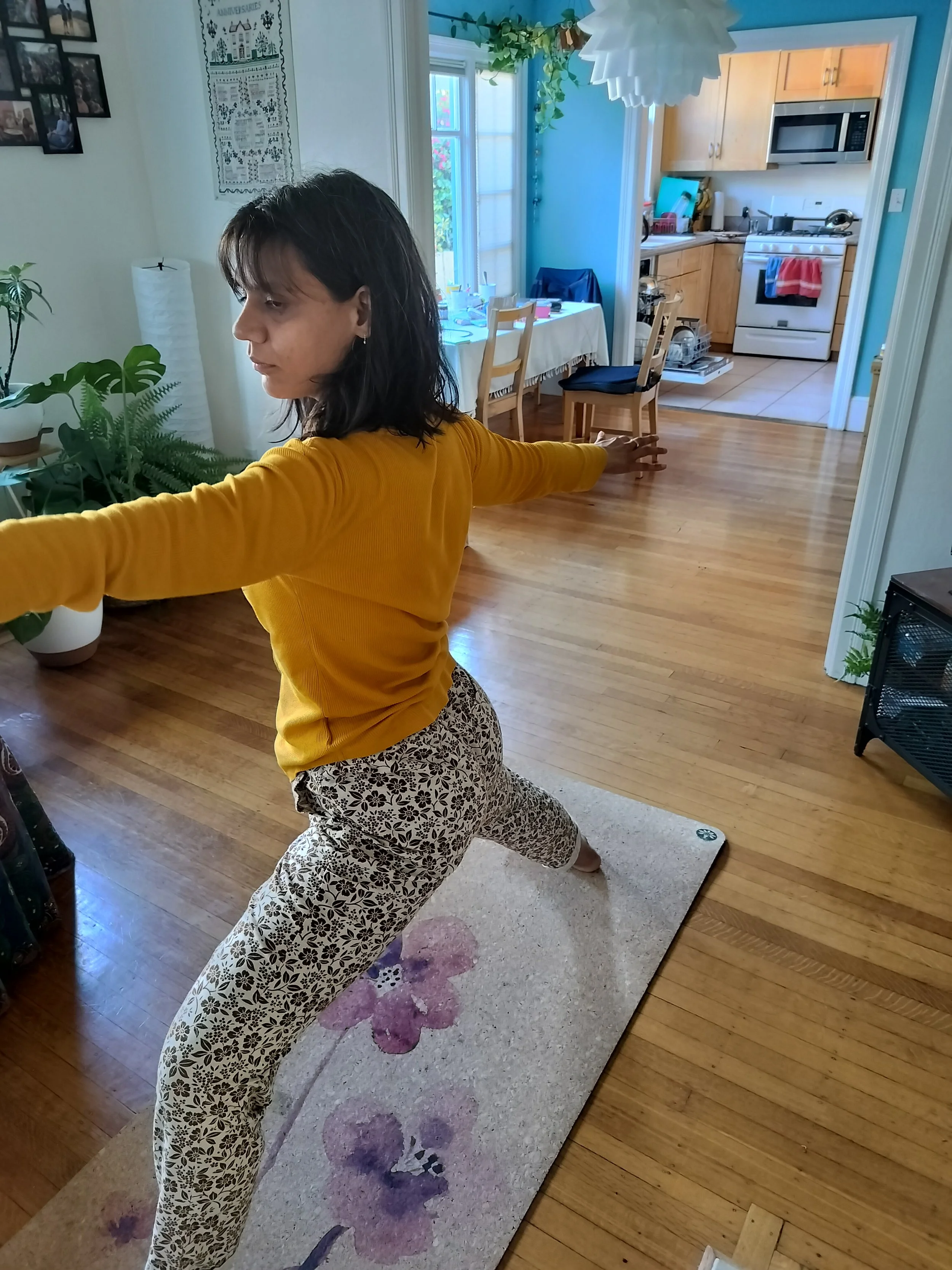 Woman practicing yoga or stretching on a floral patterned yoga mat in a living room with hardwood floors, plants, and a glimpse into a kitchen.