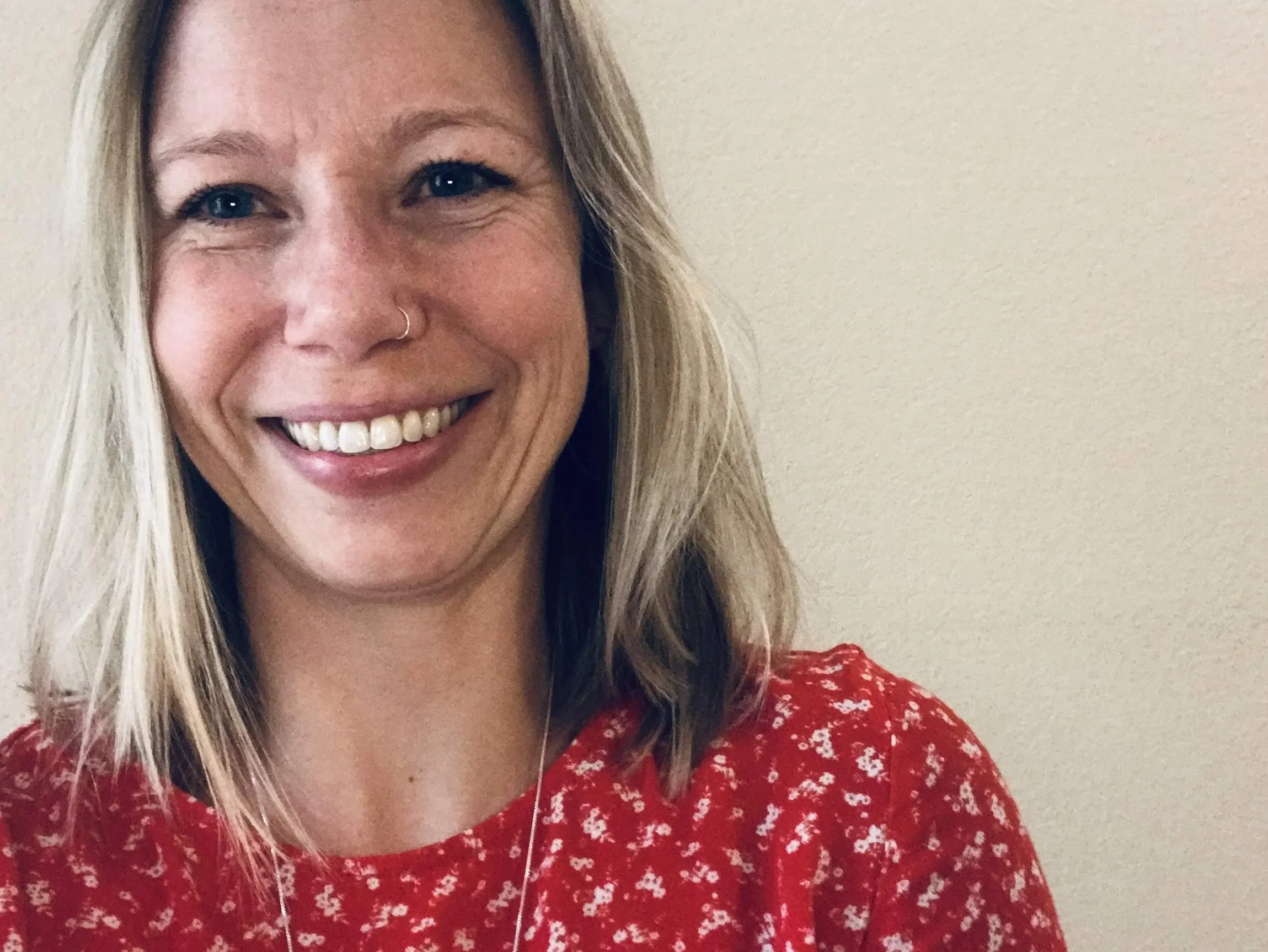 Close-up of a smiling woman with blonde hair, wearing a red patterned top and a nose ring, with a beige wall in the background.