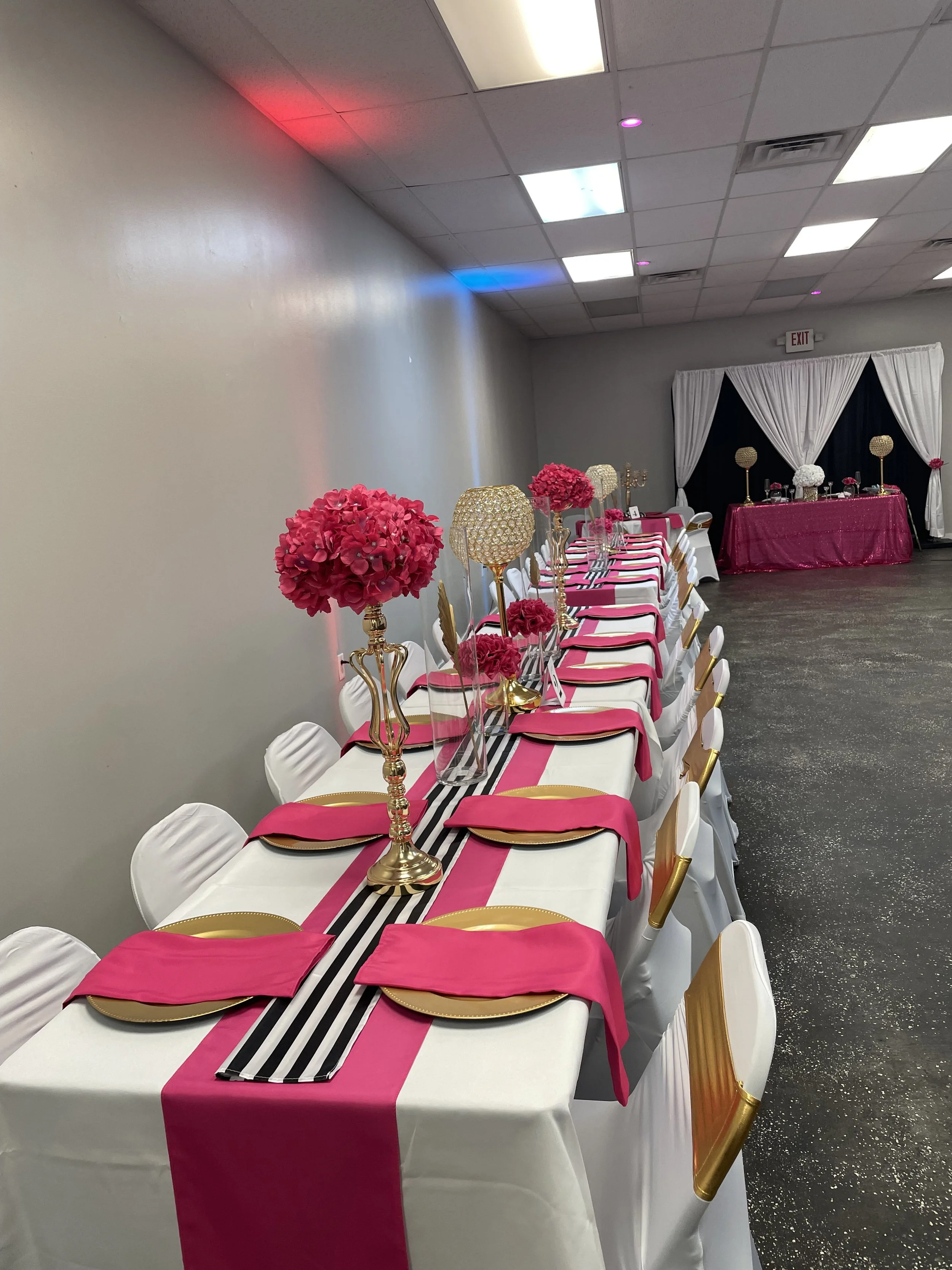 Elegant banquet table decorated with pink, black and white accents, featuring pink flower centerpieces and gold accents, set in a decorated event hall.
