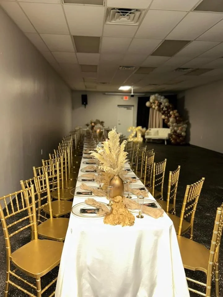 Long banquet table decorated with gold accents, beige drapes, and a large vase with cream-colored pampas grass in the center, set for a celebration or event, with golden chairs on each side and a balloon arch at the back.
