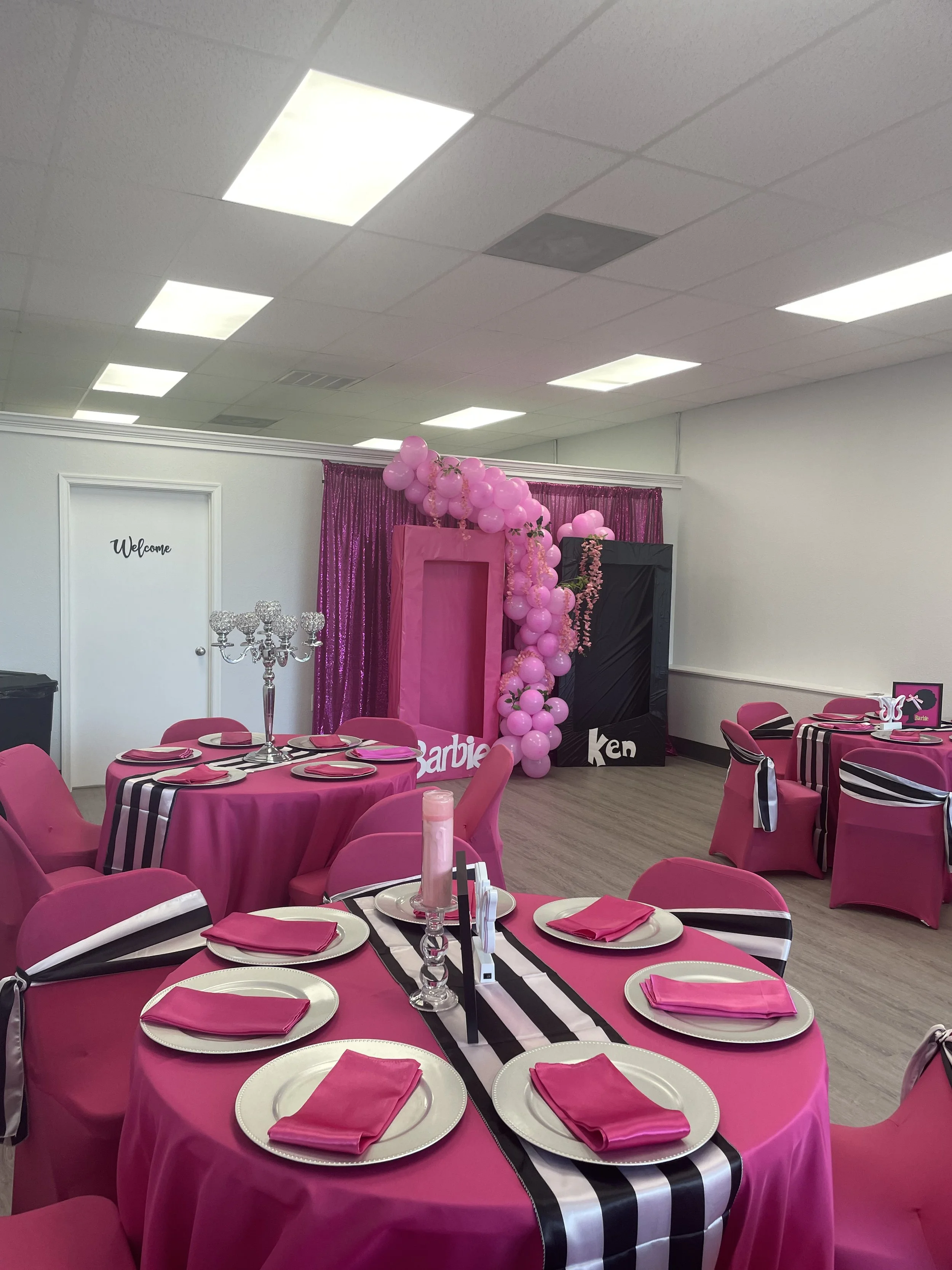 Decorated party room with pink tablecloths, pink chairs, a pink and black striped runner, pink napkins, a silver candelabrum, and a backdrop with pink balloons, a pink frame, and a black and pink display with the words 'Barbie' and 'Ken'.