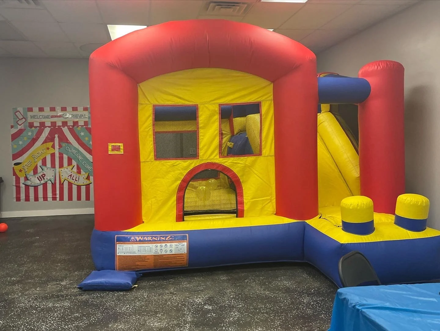Colorful indoor inflatable castle with a yellow slide, red and yellow structure, and mesh windows, set up in a room with a black speckled floor and a wall decorated with carnival-themed signs.