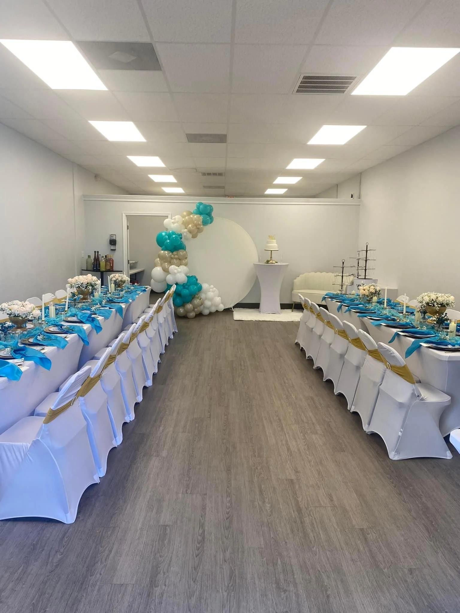 Wedding reception room with long tables decorated with white covers, gold sashes, and blue napkins. There are white flower centerpieces, a balloon arch in teal, white, and gold, a cake on a high table, and a seating area with a white couch and a smal