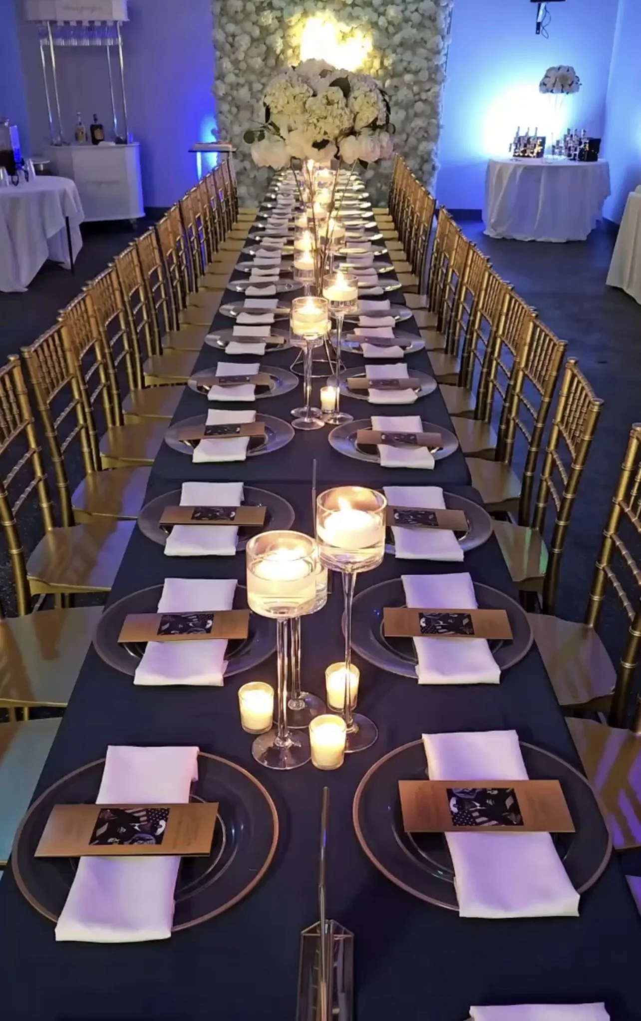Elegant banquet table decorated with candles, tall floral centerpieces, and place settings for a formal event, in a dimly lit room with a decorated backdrop.