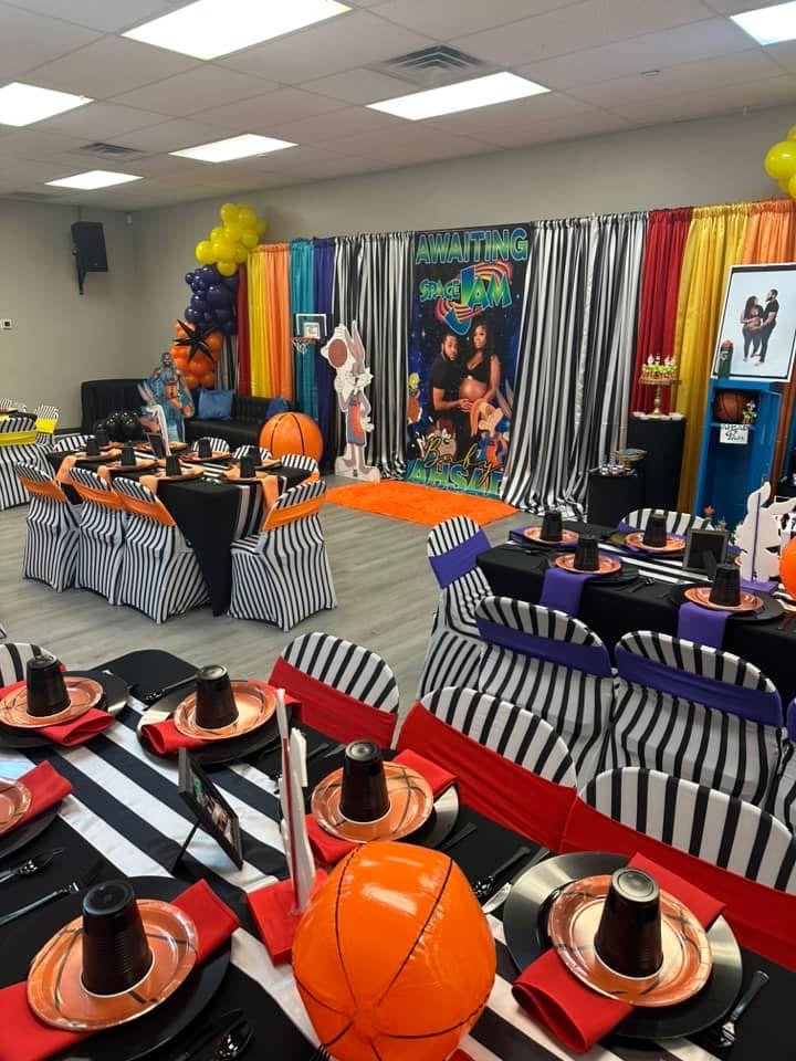 Party room decorated with black and white striped tablecloths, orange, purple, and red accents, bat-themed decorations, a stage with a backdrop of colorful curtains, and themed posters and balloons, suggesting a Halloween or spooky-themed celebration.
