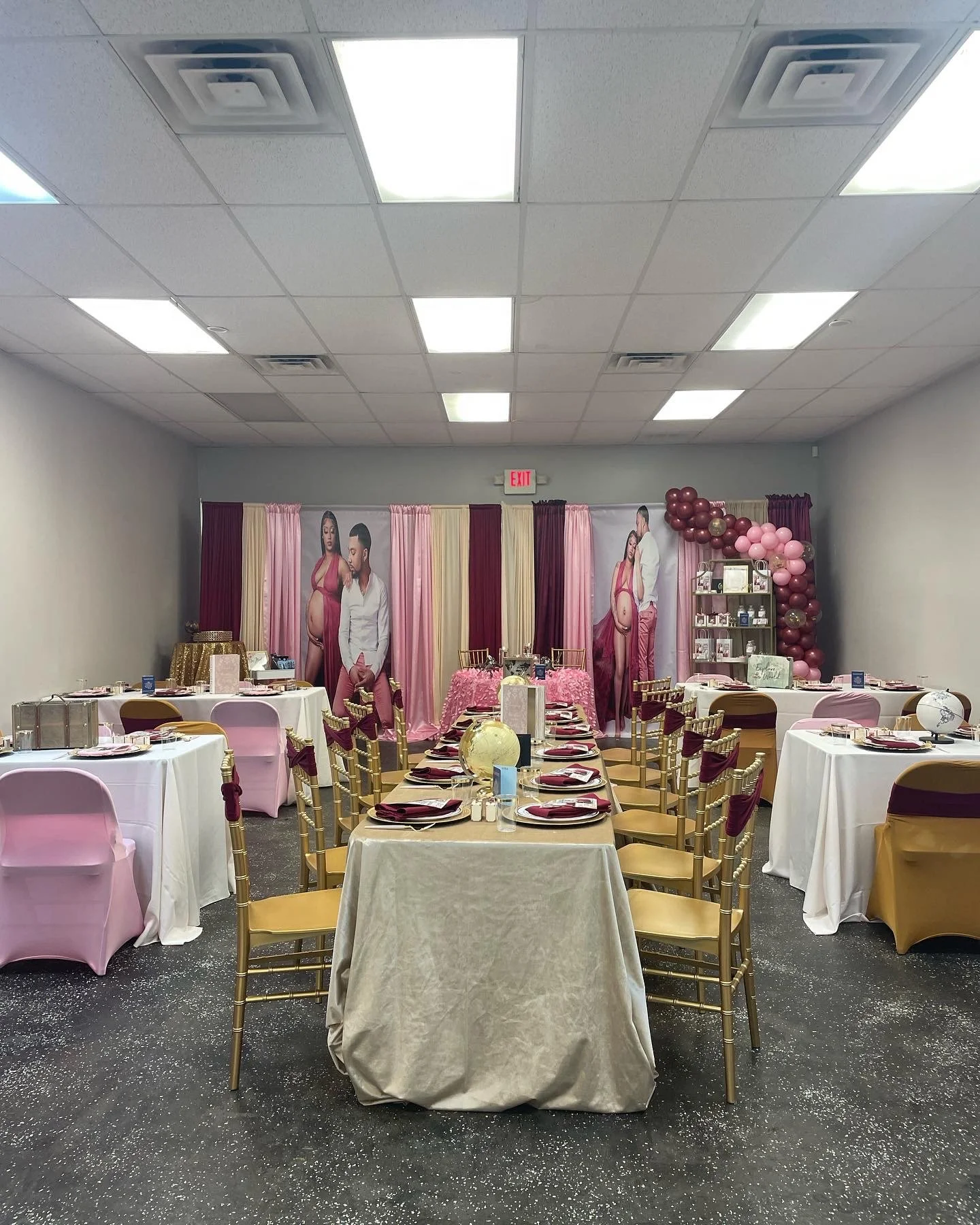 Baby shower decorated with pink, gold, and maroon colors. Tables with place settings, balloons, and a large backdrop with images of an expecting couple.